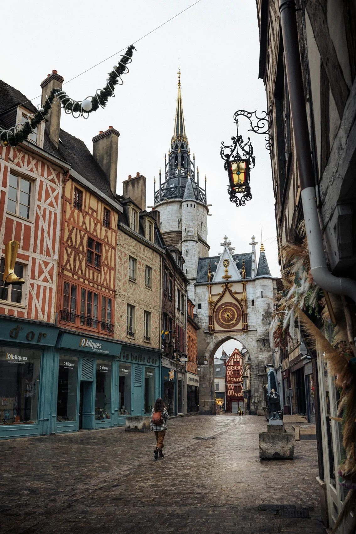 Rue pavée avec des bâtiments à colombages, une horloge médiévale, au sommet d'une tour, et un clocher, avec des décorations de Noël suspendues.
