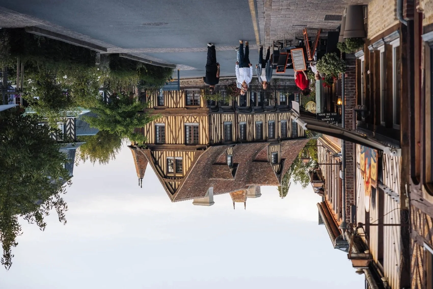 Rue pavée avec des bâtiments en style traditionnel et des terrasses de cafés, arbres verdoyants et ciel clair.