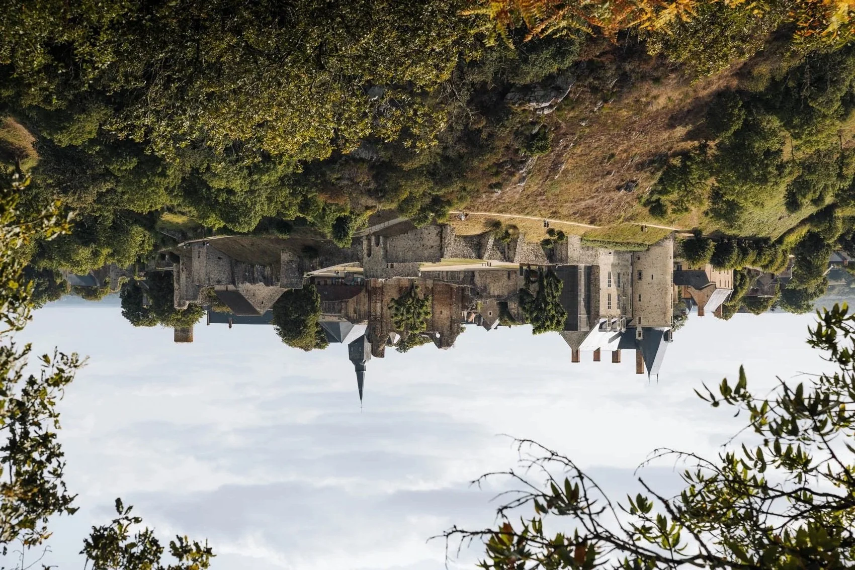 Château médiéval en pierre au sommet d'une colline, entouré d'arbres, avec un ciel nuageux en arrière-plan.