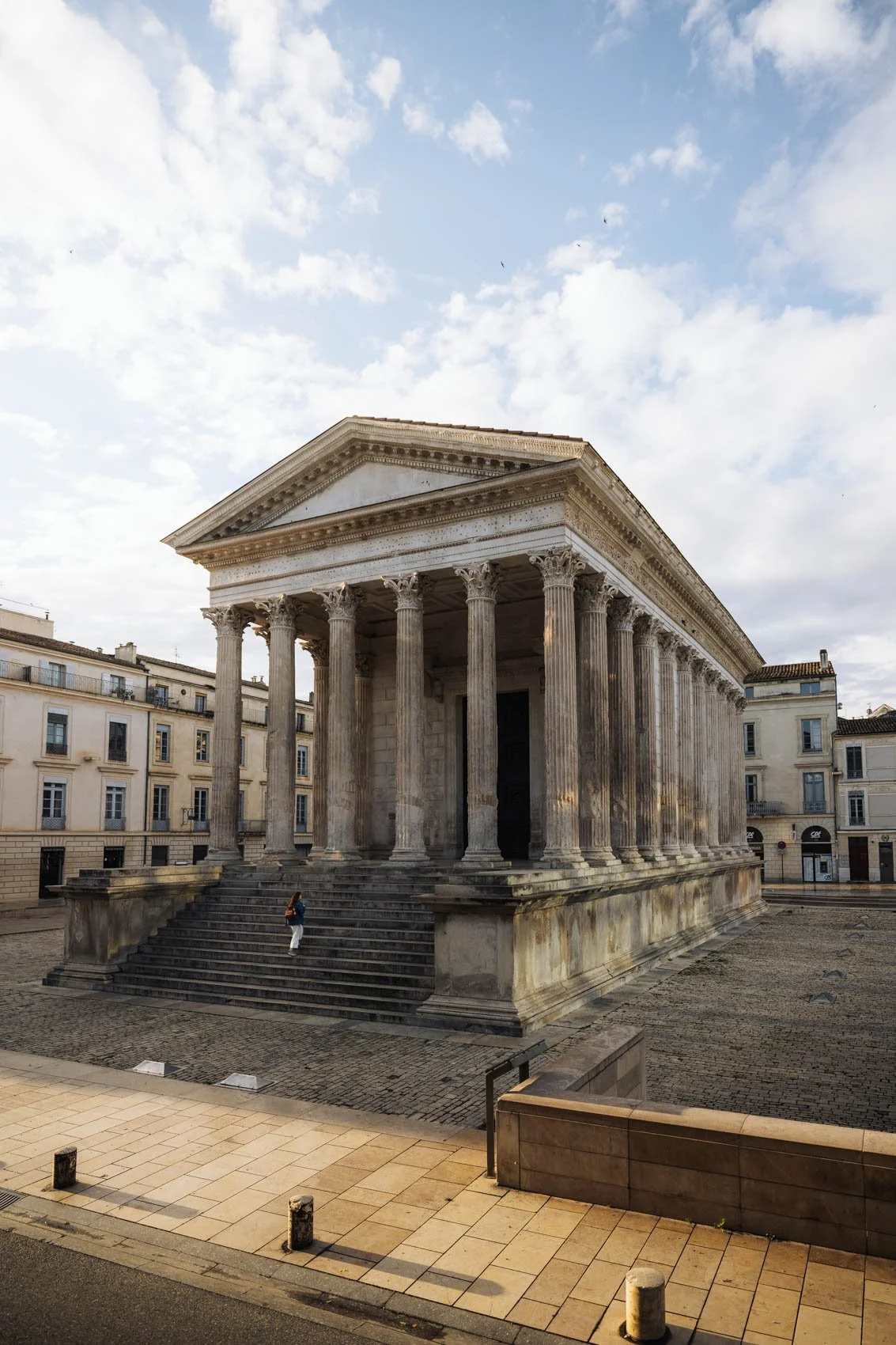 Un bâtiment en style grec antique avec des colonnes et des escaliers en pierre, situé dans une place pavée. Le ciel est partiellement nuageux.