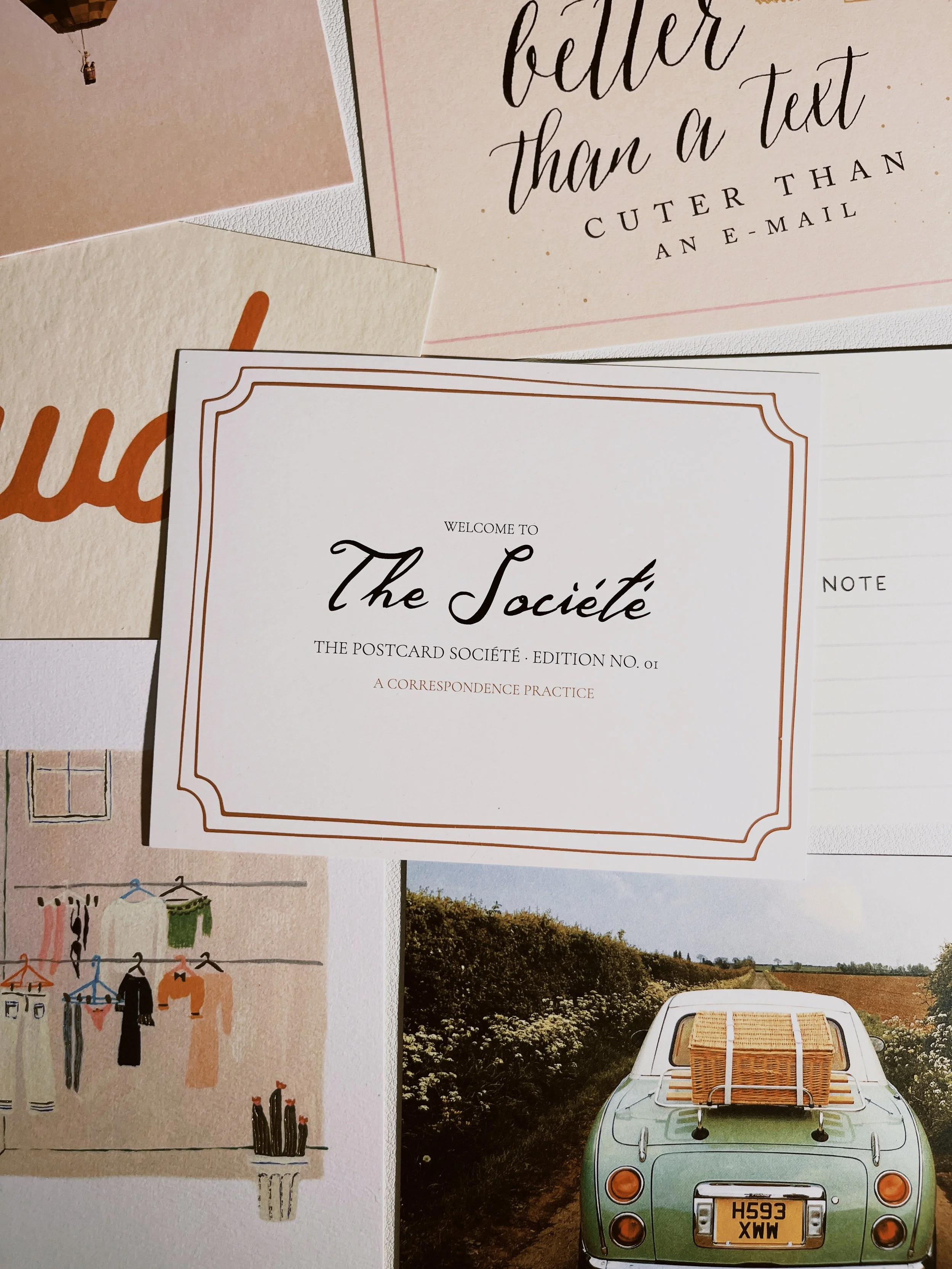 Postcards scattered on a desk with a welcome card on top designed for Edition 01 of The Postcard Société, postcard correspondence subscription box curated by social and cultural psychologist and travel psychologist Dr. Katie Blake, PhD.