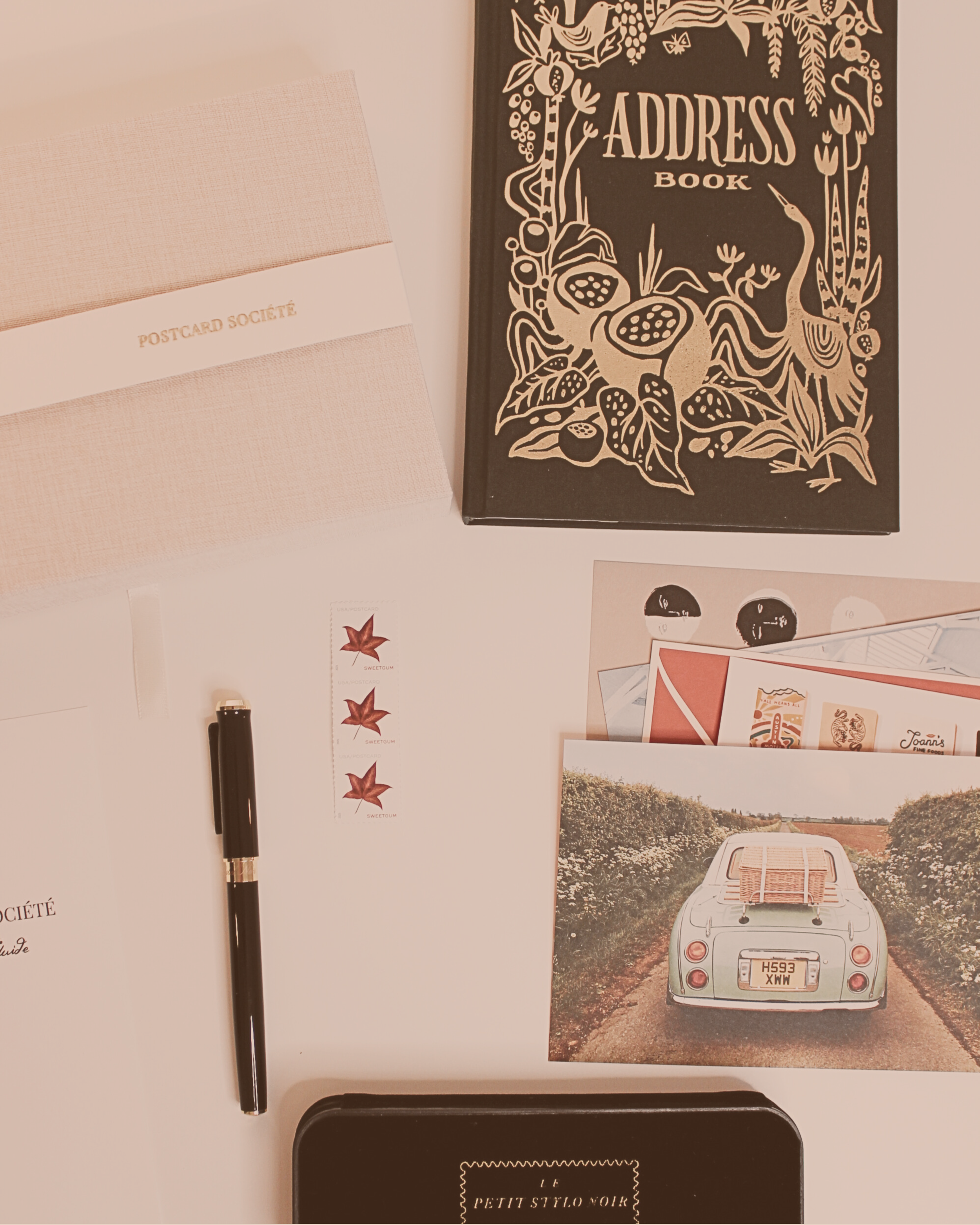 A flat lay of The Postcard Société Edition 01 contents arranged on a soft blush surface — a linen keepsake box embossed with "Postcard Société," a cloth-bound address book with gold illustrated cover, a black correspondence pen and its case, three re
