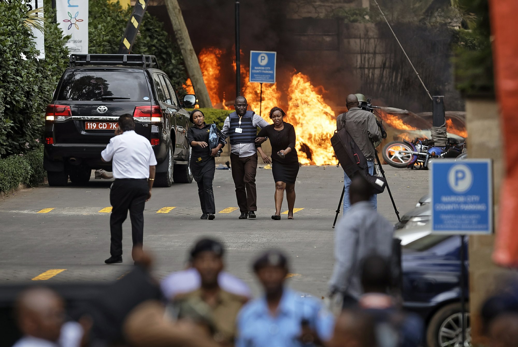 Security forces help civilians flee the scene as cars burn behind, at a hotel complex in Nairobi, Kenya Tuesday, Jan. 15, 2019. Terrorists attacked an upscale hotel complex in Kenya's capital Tuesday, sending people fleeing in panic as explosions and heavy gunfire reverberated through the neighborhood. (AP Photo/Ben Curtis)