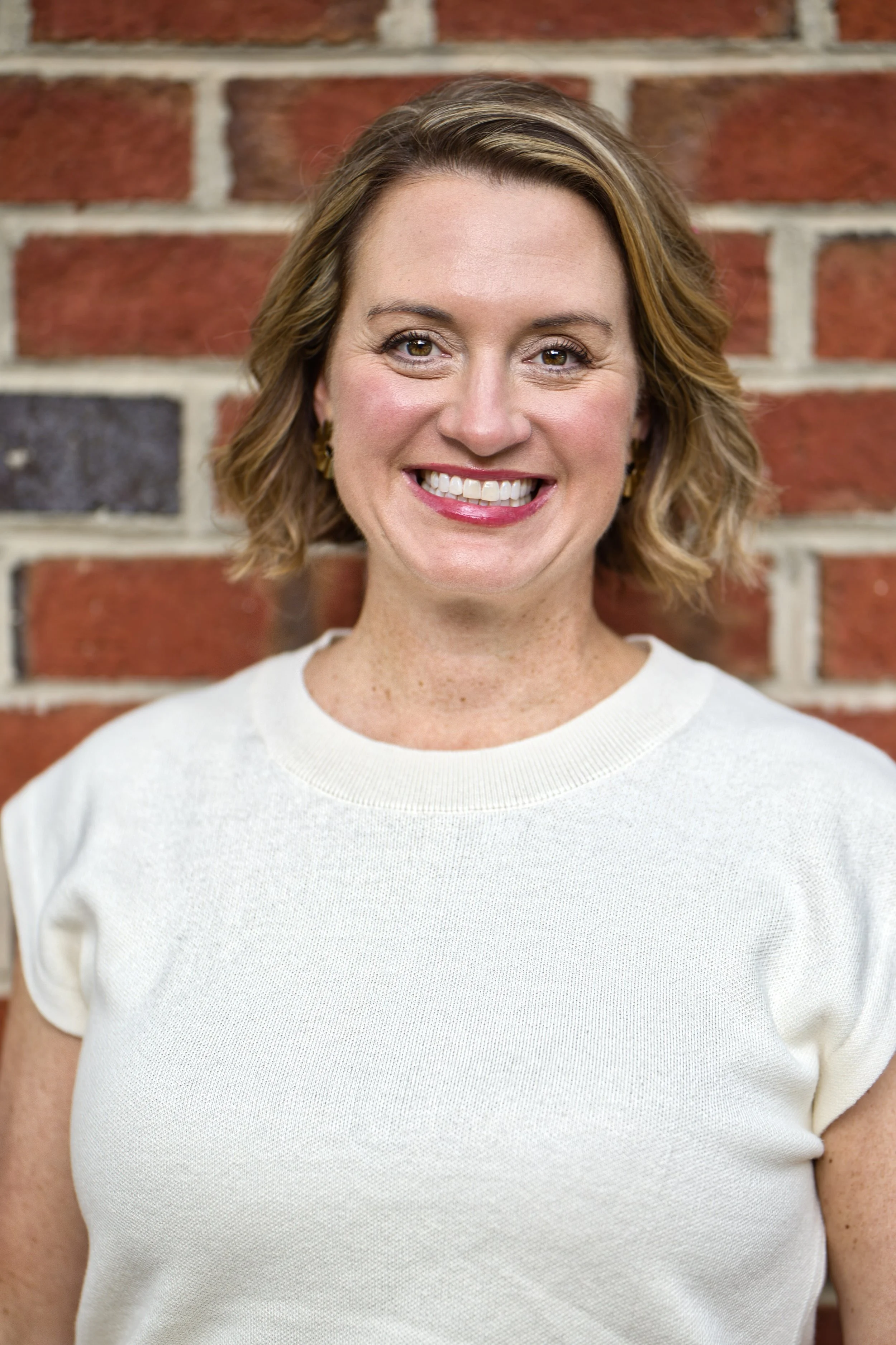 Close-up of a smiling woman with short, wavy brown hair, wearing a white sweater, standing against a brick wall background.