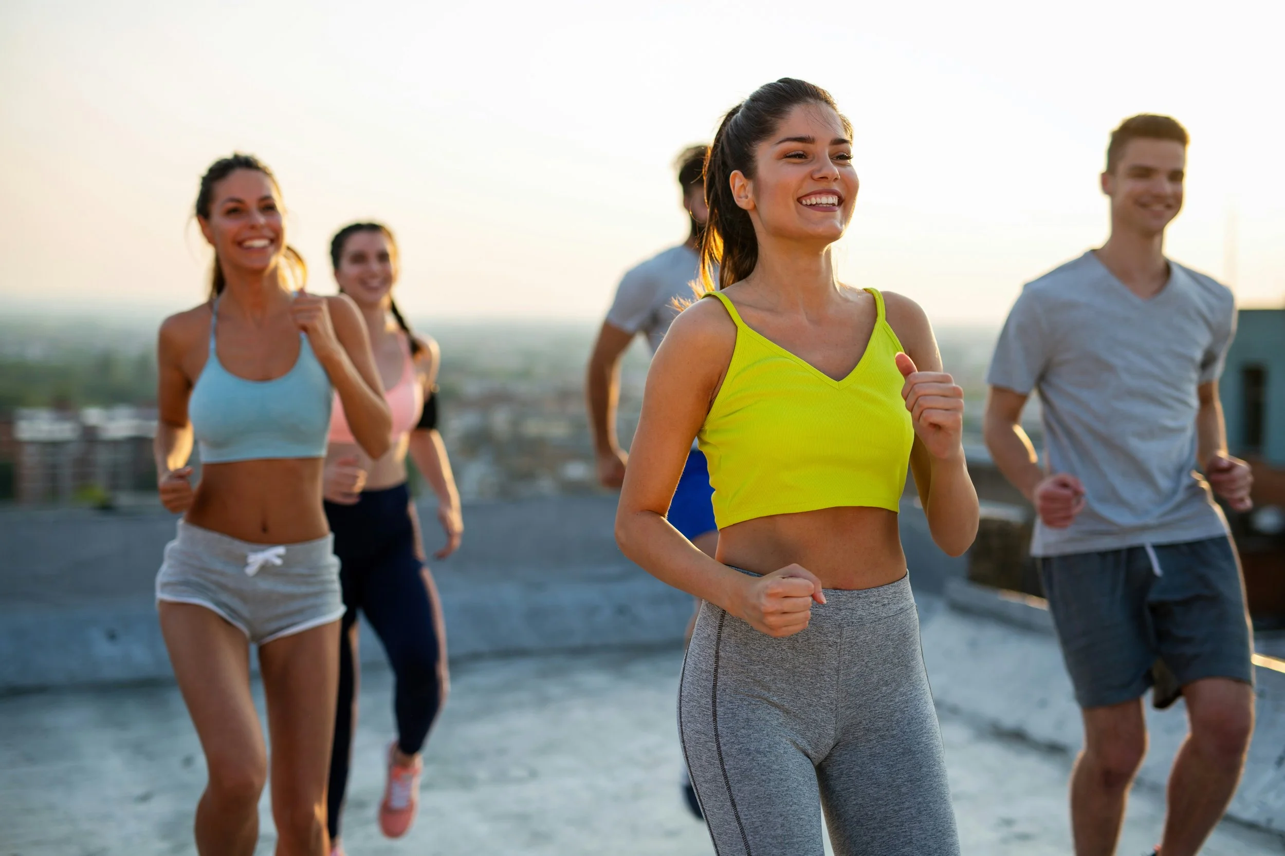 Group of five people running outdoors on a rooftop during sunset, smiling and enjoying exercise.