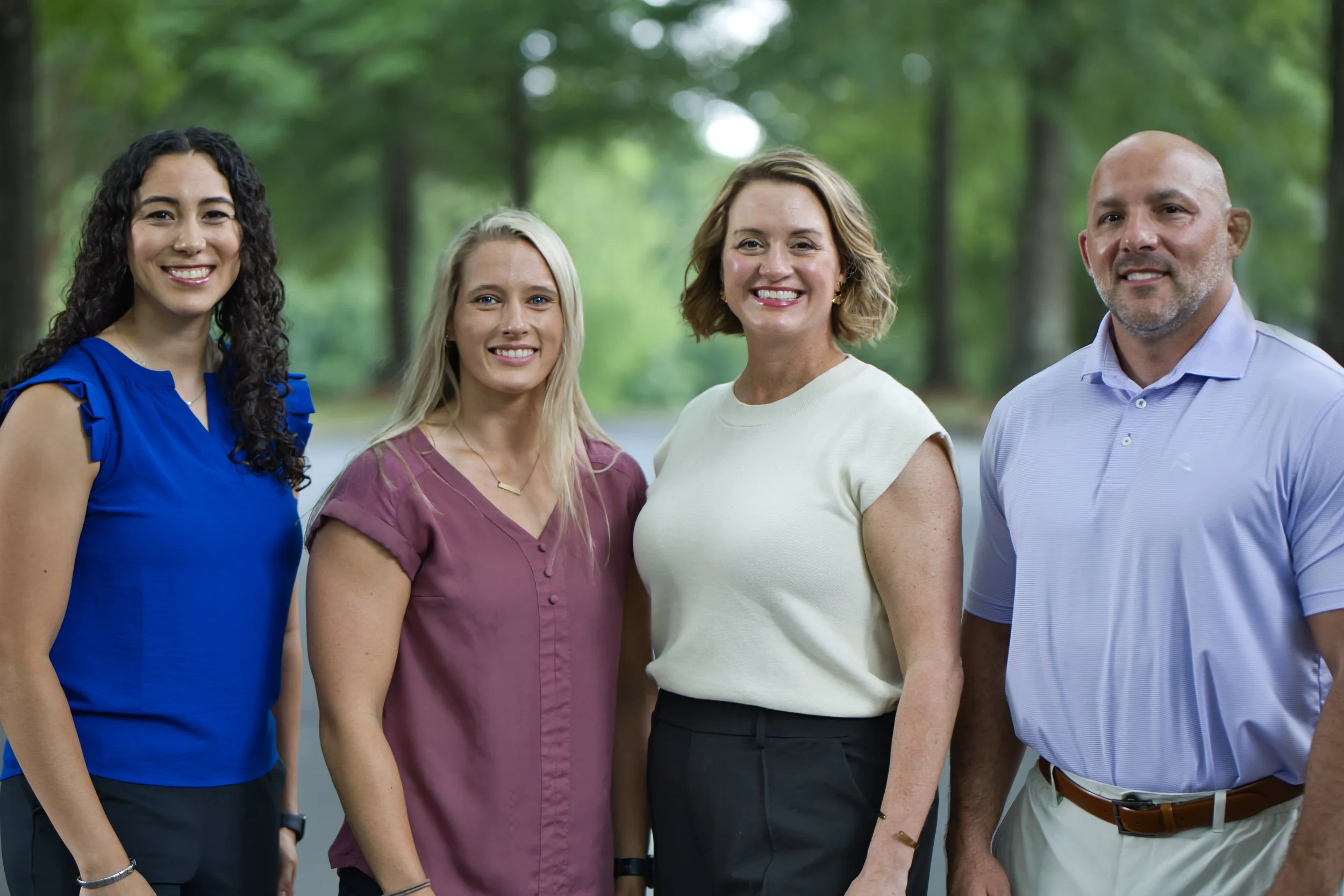 Four diverse adults standing outdoors in a green, wooded area, smiling at the camera.