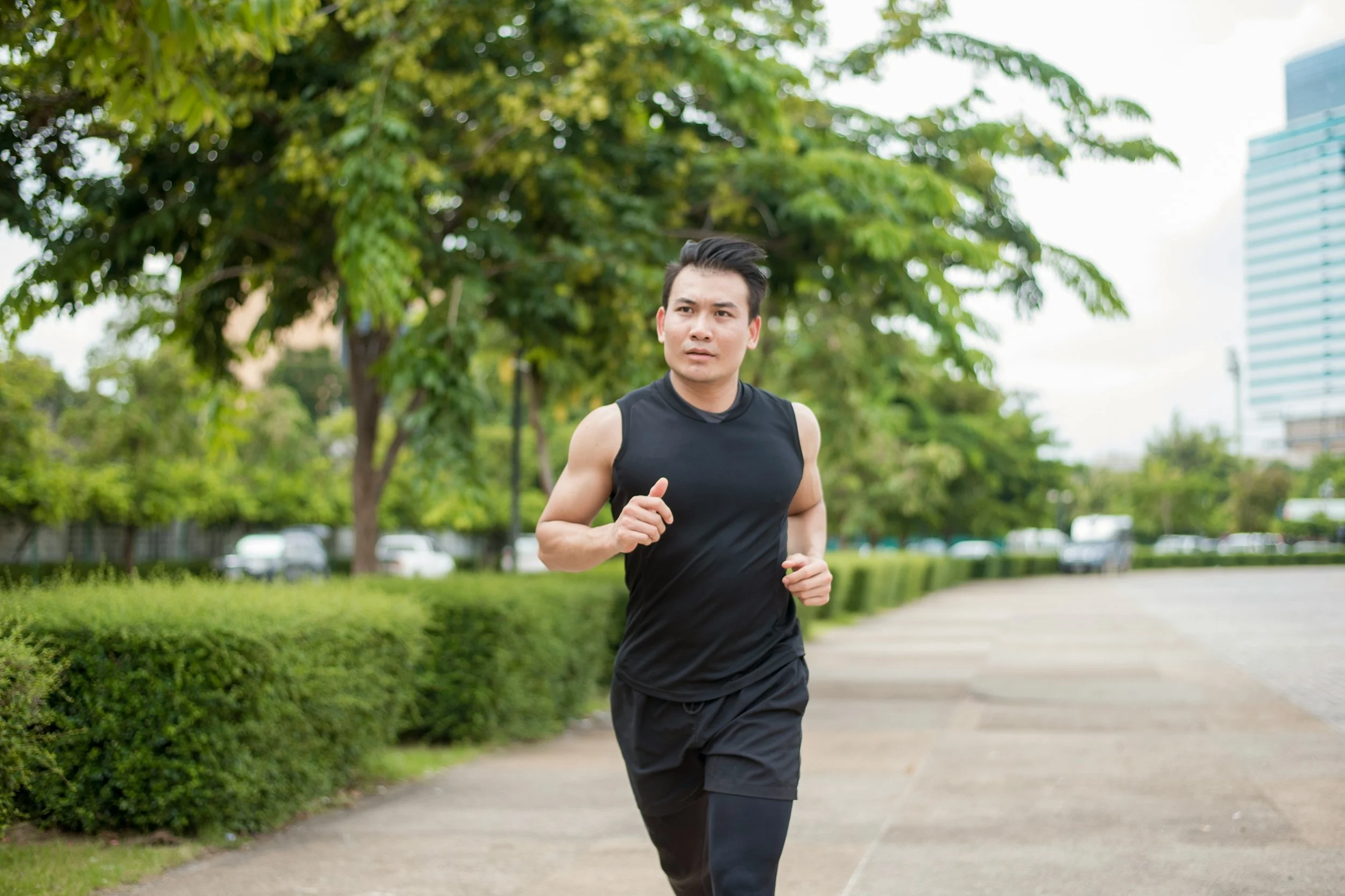 A man running outdoors on a paved pathway surrounded by green trees and bushes, with buildings and cars in the background.