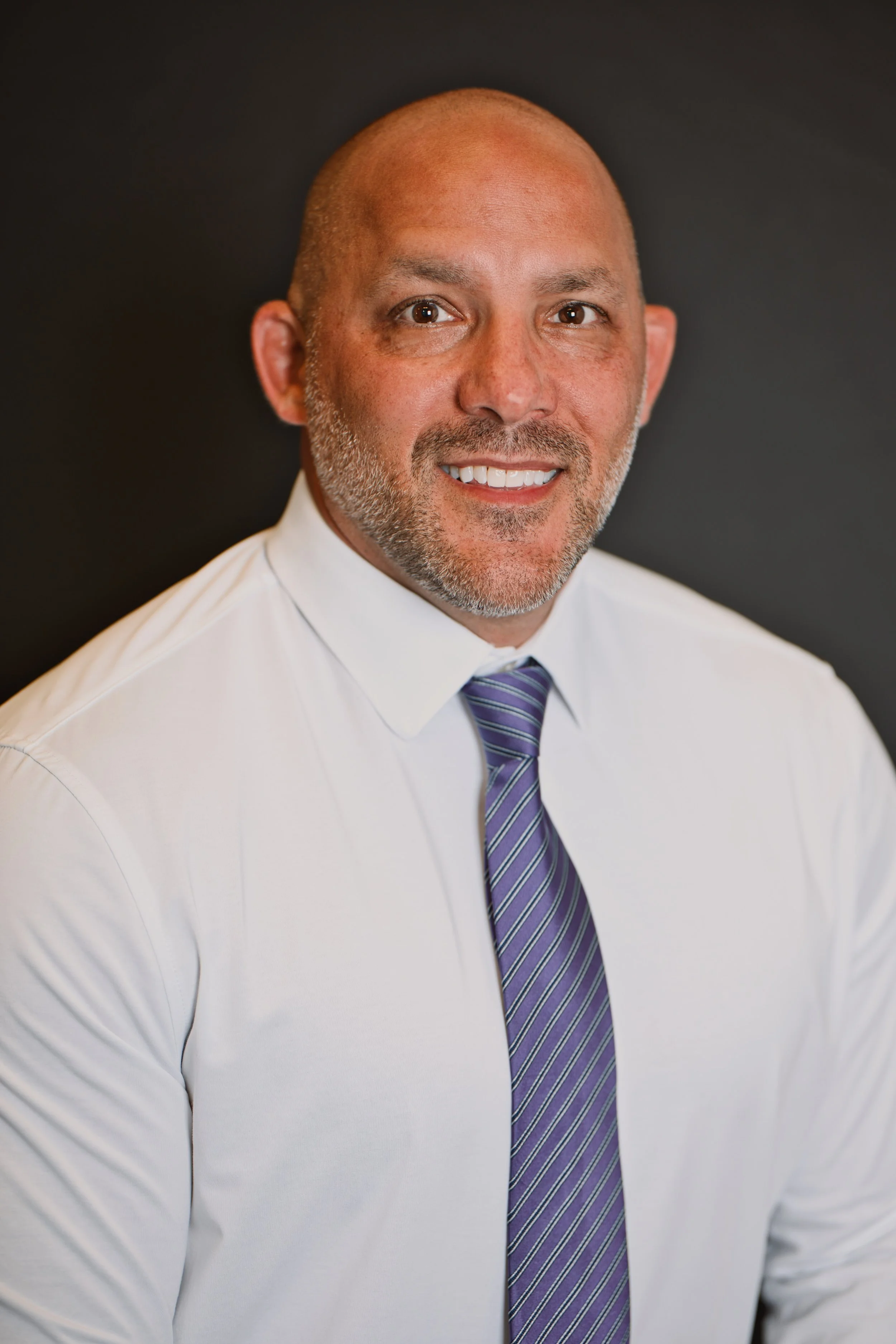 Headshot of a good looking smiling man with a salt and pepper beard, wearing a white shirt and a purple striped tie, against a dark background.