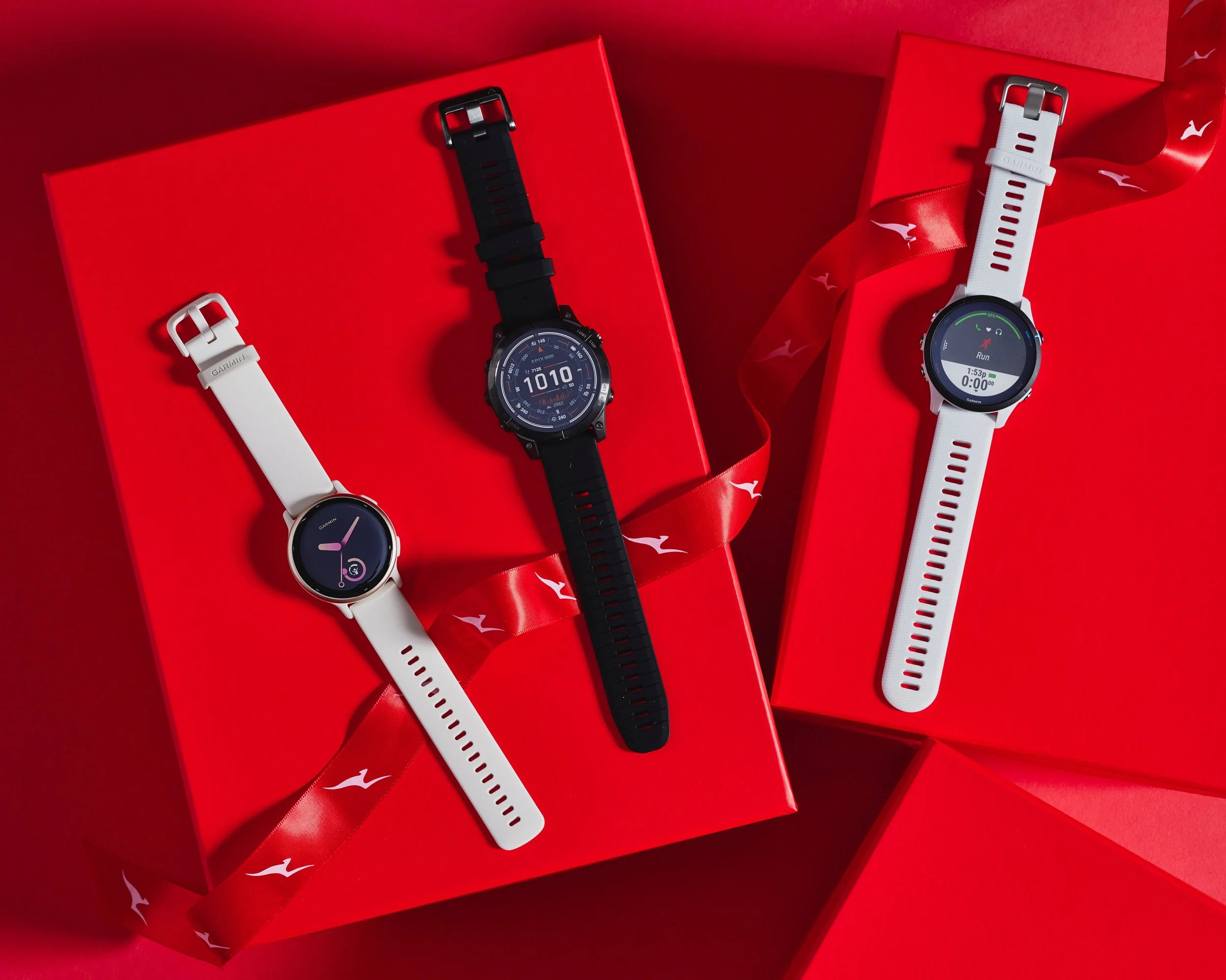 Two Garmin smartwatches, one with a white strap displaying activity data and the other with a black strap showing a temperature of 101°F, rest on red gift boxes with white and red ribbon accents.
