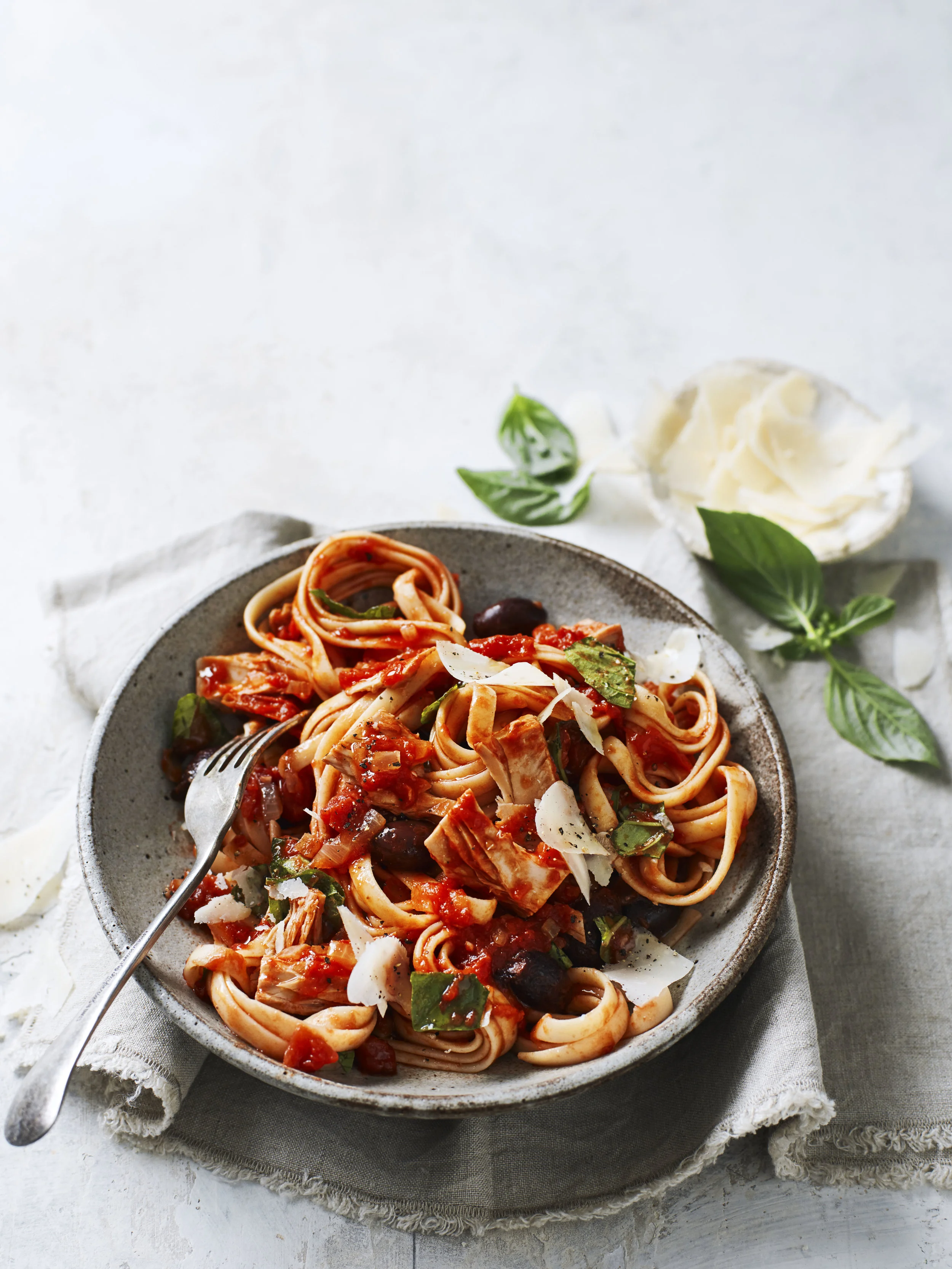 A bowl of spaghetti with tomato sauce, garnished with basil, served on a piece of cloth with a fork. Garnished cheese and basil leaves on the side.