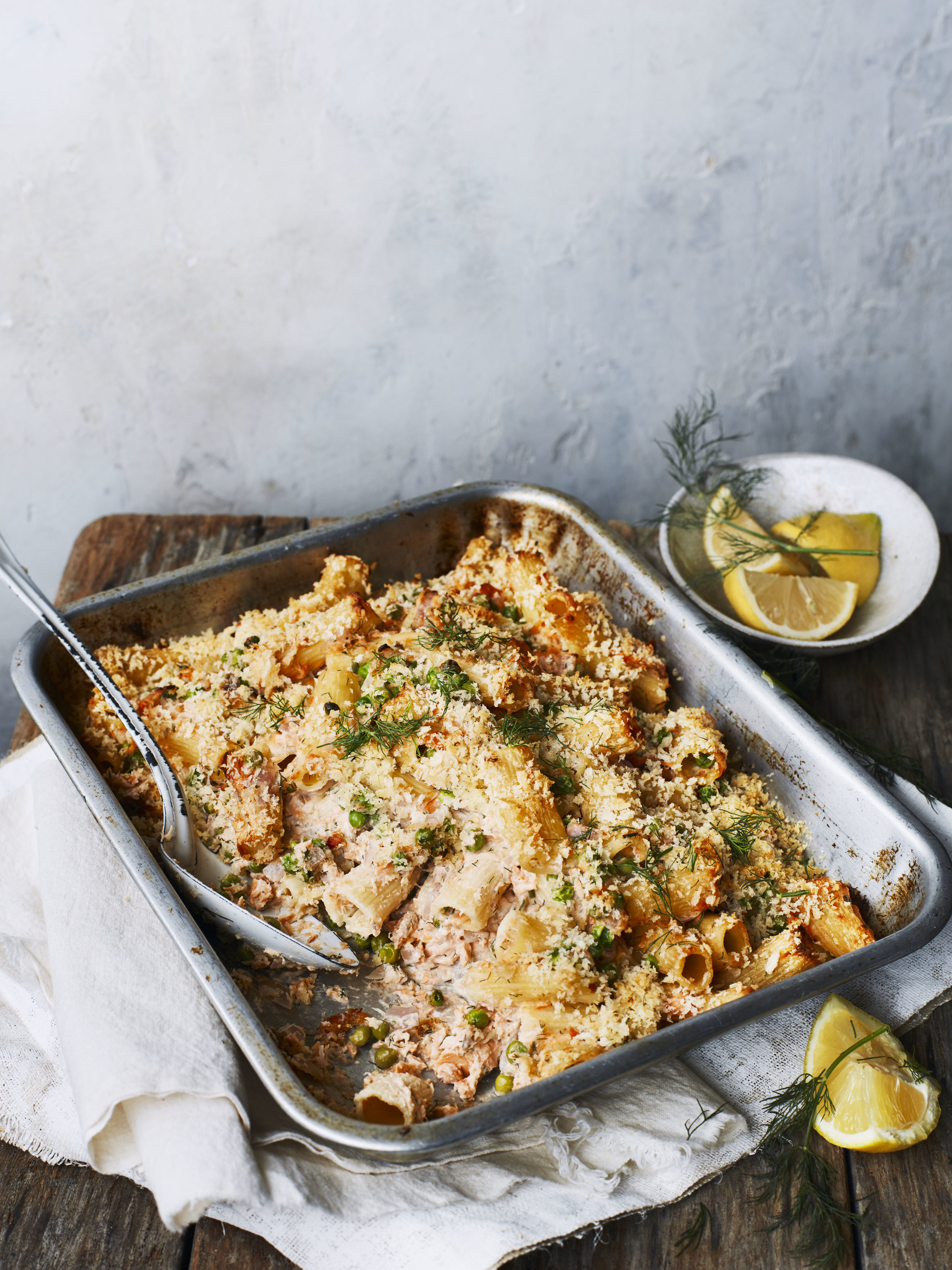 Baked pasta casserole in a metal dish garnished with herbs, with lemon wedges and dill on the side, on a wooden surface.