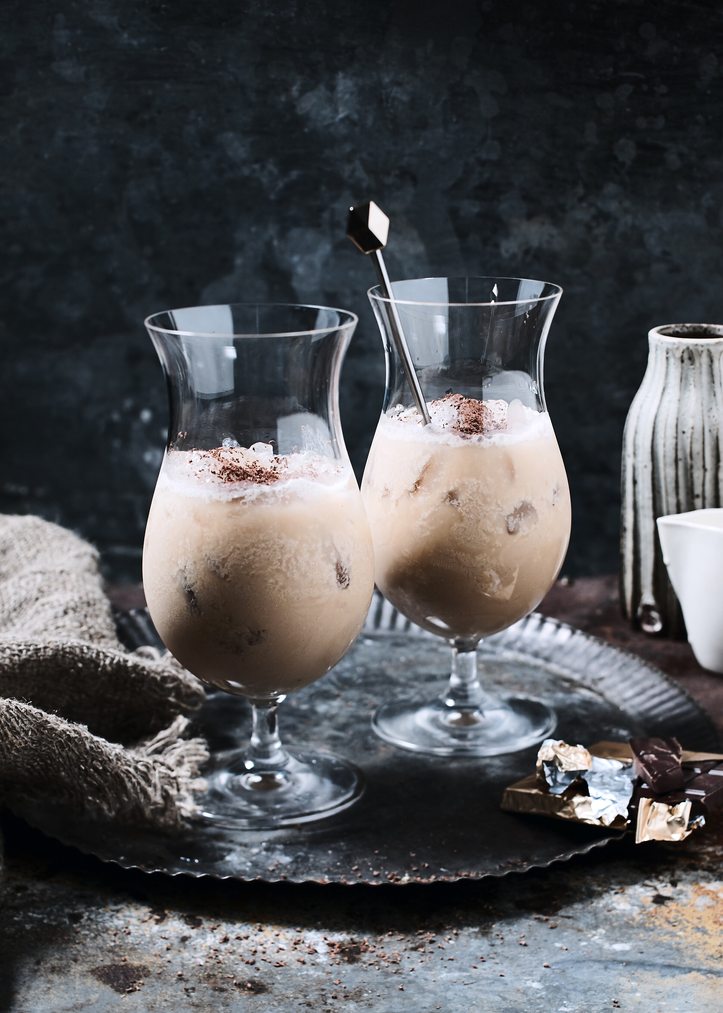 Two glasses of frothy chocolate milkshake topped with cocoa powder, set on a dark surface with scattered chocolate pieces, a wrapped chocolate bar, and a pottery vase in the background.