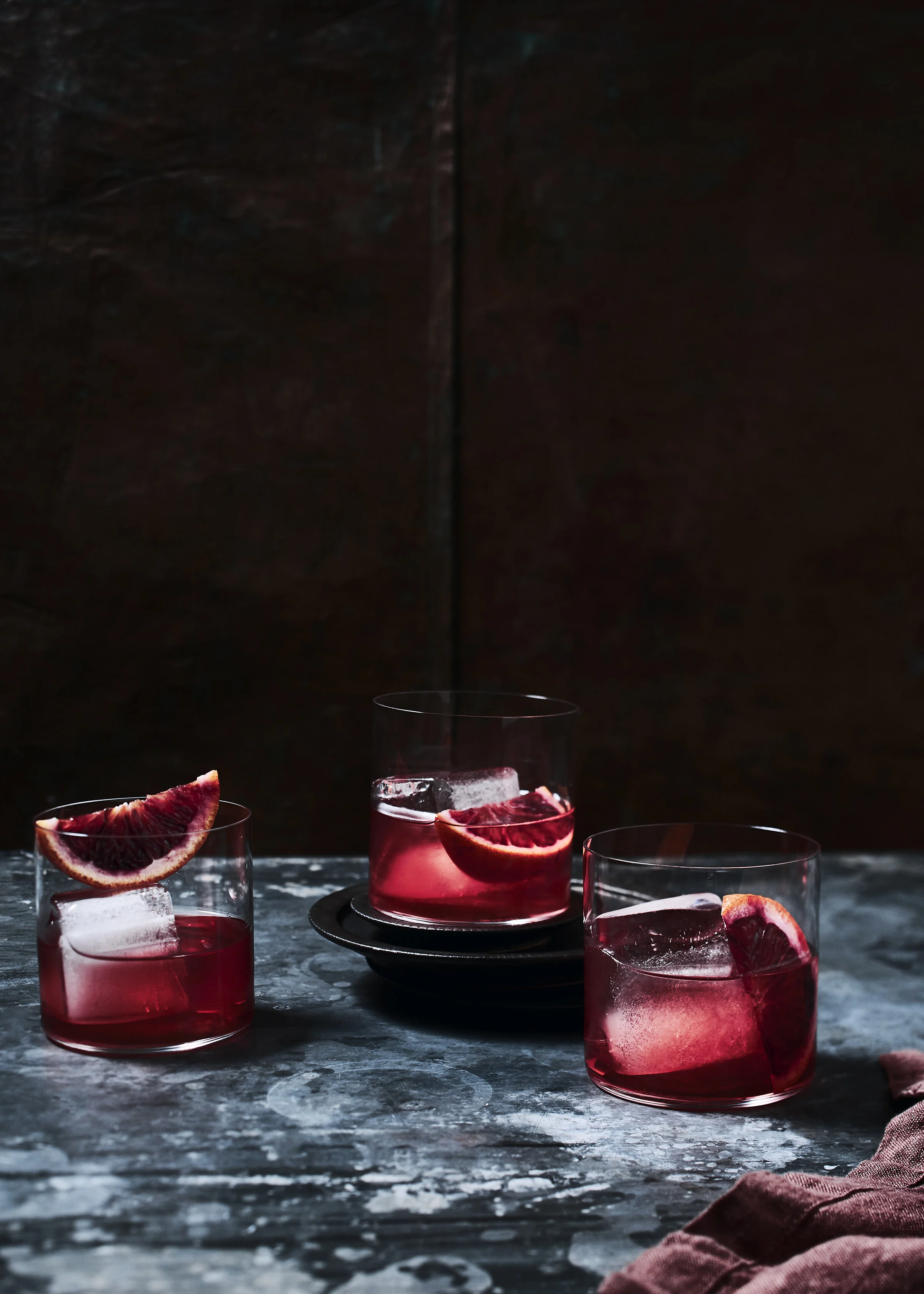 Four glasses of pink-hued beverage with ice and slices of grapefruit on top, placed on a dark textured surface with a dark background.
