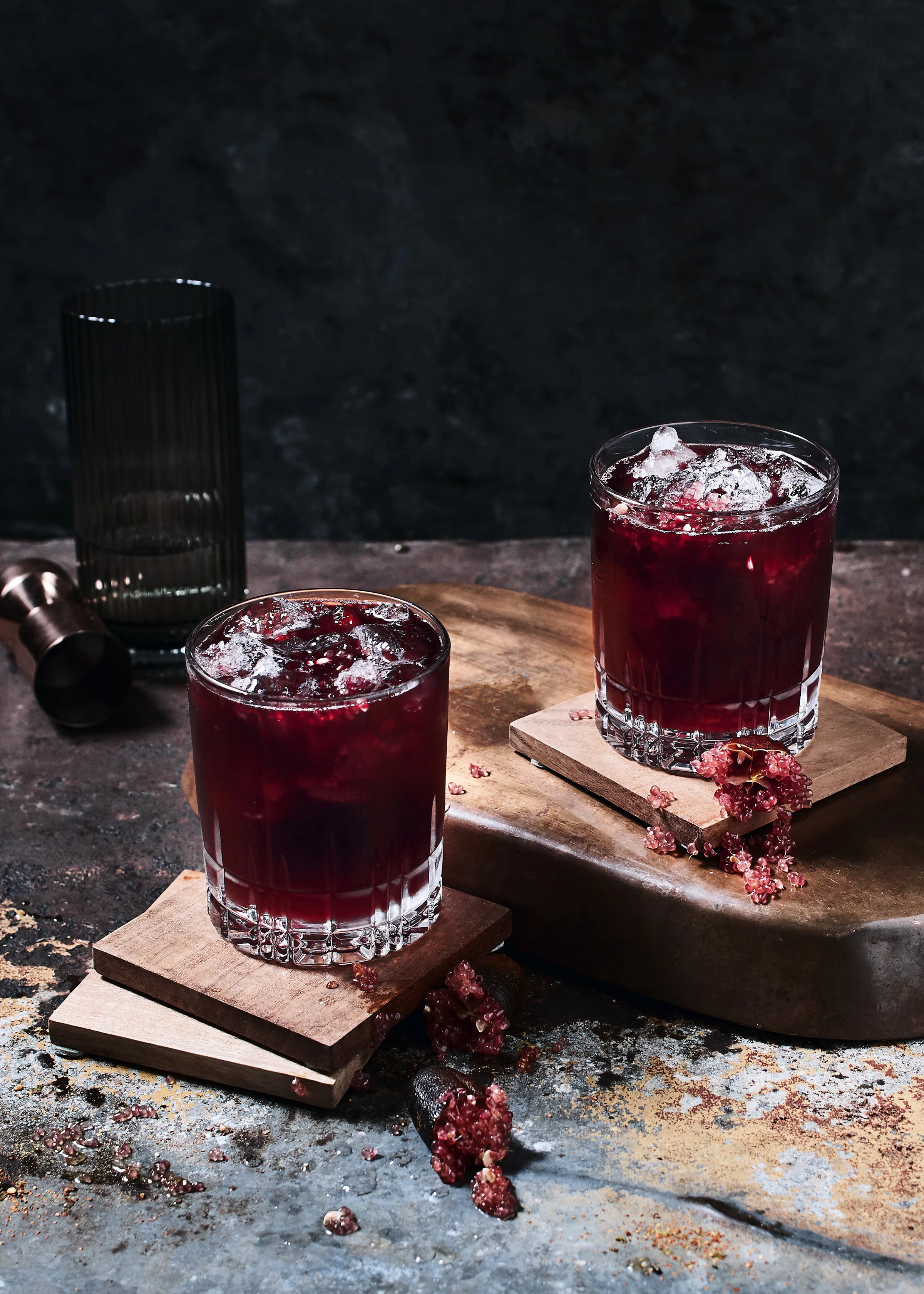 Two glasses of red beverage with ice on a wooden surface, with a bunch of pink berries and spilled berries around, and a black glass and metal object in the background, dark textured surface behind.