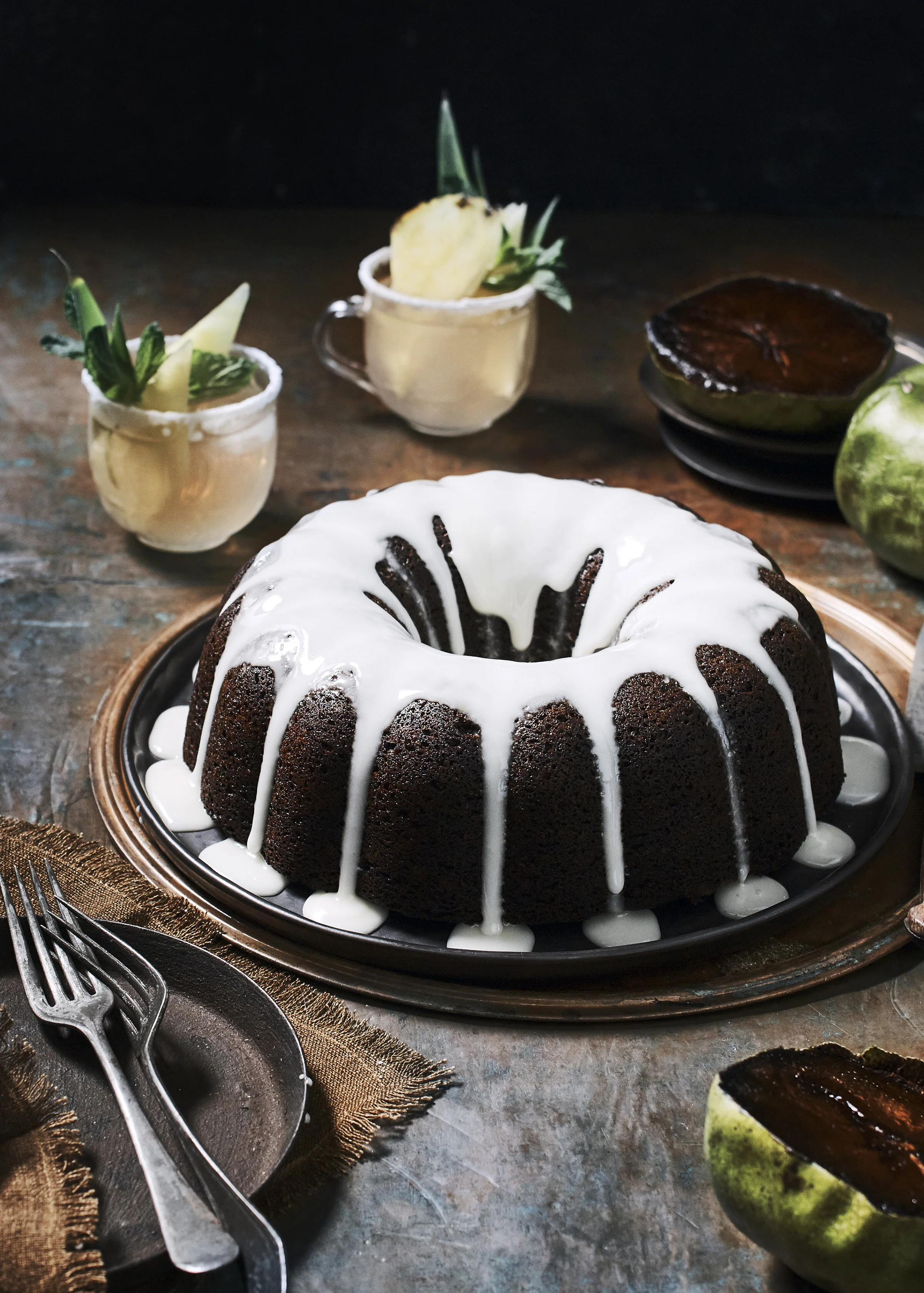 Chocolate bundt cake with white icing and slices of pineapple garnished with green leaves, set on a copper-colored tray on a rustic table, surrounded by unevenly stacked plates with caramelized tops and halved green apples with caramelized filling.