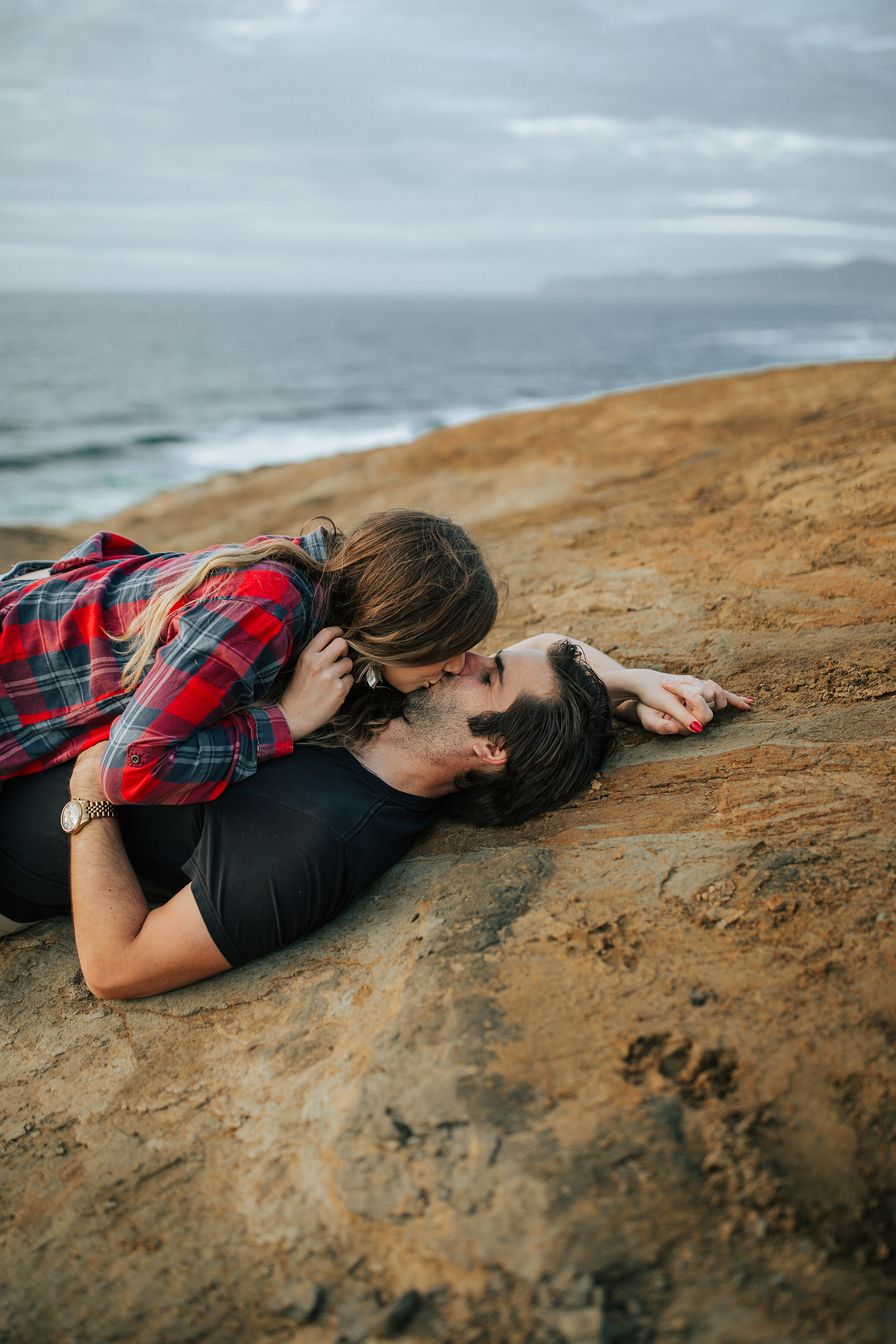  A beautiful couple share a romantic kiss by the stormy ocean in a dark and romantic couples honeymoon photo shoot by Emily Jenkins Photography. Laying down couples pose inspiration ideas and goals kissing couple pose inspiration client attire inspir