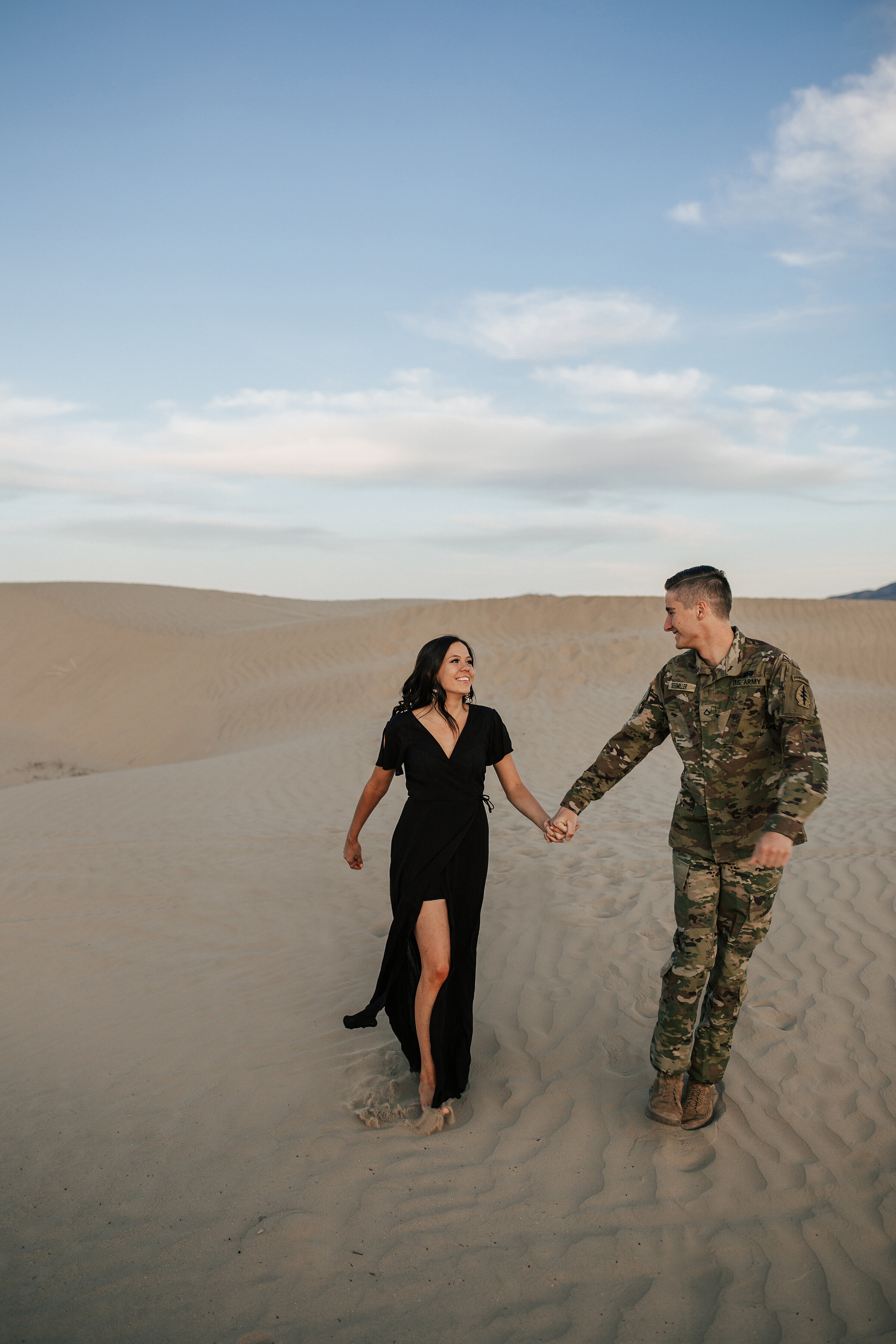  A service man and his wife walk through the sand dunes in a stunning engagement session by Emily Jenkins Photography. Professional Utah photographer engagement photo shoot semi formal engagement outfit inspiration  outdoor photo shoot inspiration se
