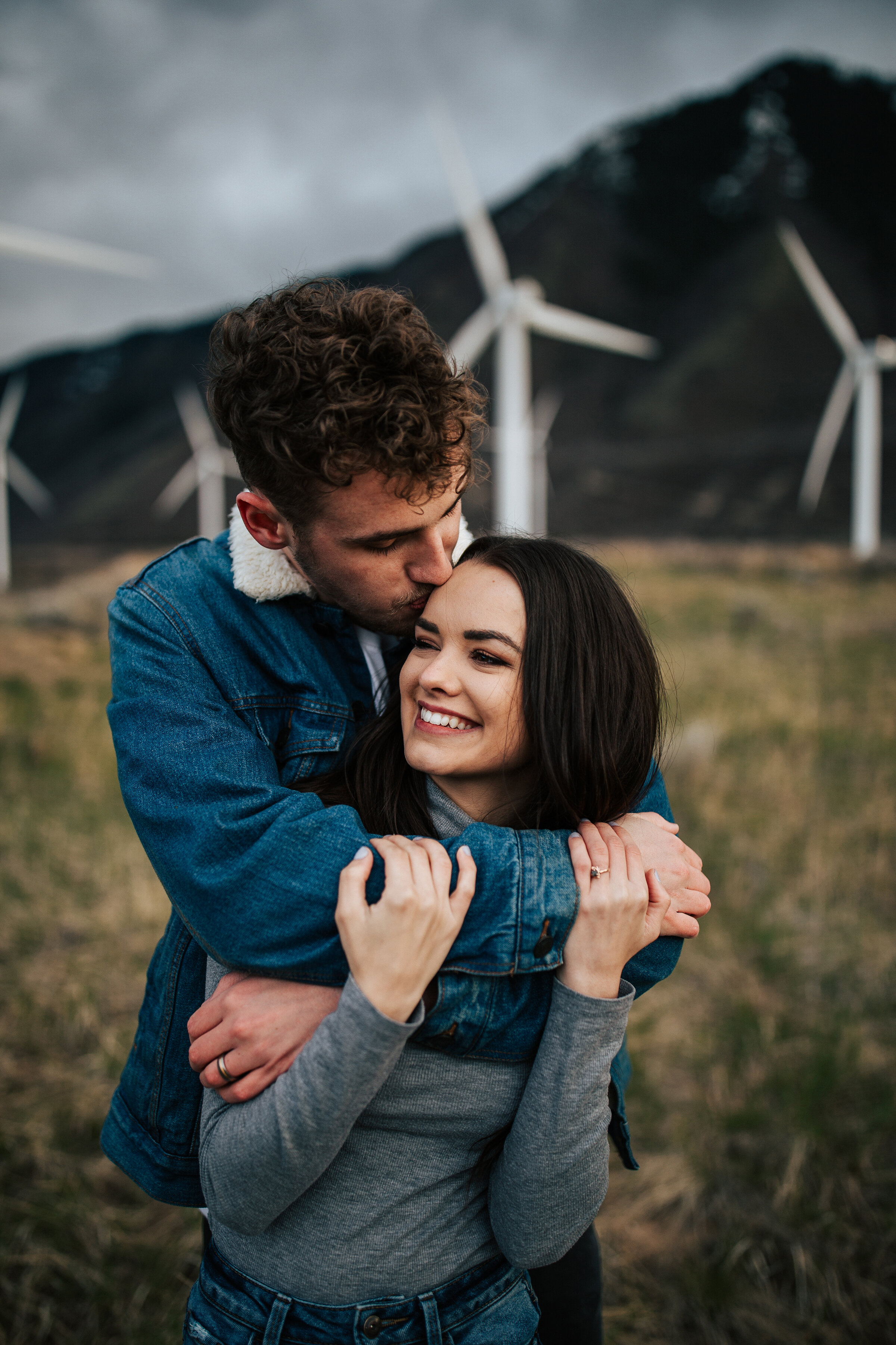  A man kisses his stunning fiancés forehead as they stand in a windmill field  in a unique engagement session  by professional engagement photographer Emily Jenkins Photography. Couple pose inspiration casual engagement session outfit inspiration for