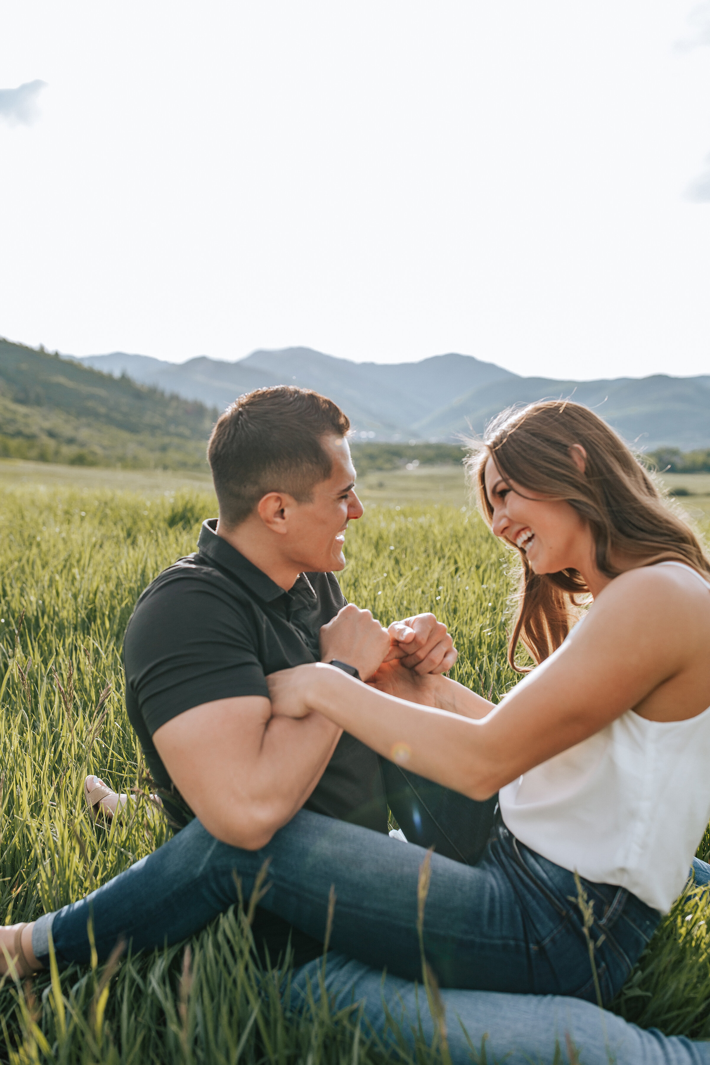  Laughing couple sit together in a grassy field in a beautiful summer engagement session by professional Utah photographer Emily Jenkins Photography. Summer or spring engagement session inspiration ideas and goals sitting couple pose inspiration casu