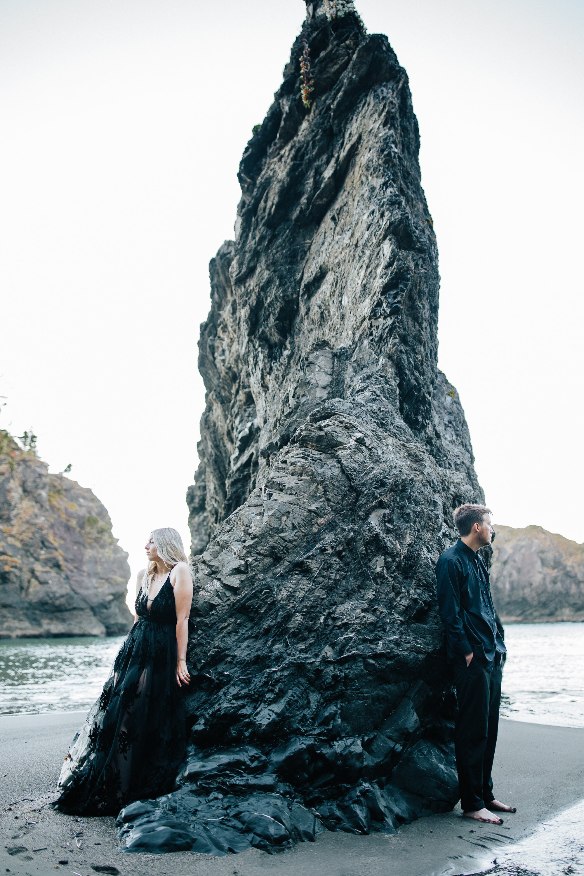  A couple wearing black during a beach elopement in Brookings, Oregon stand with their backs against a black pillar cliff by Emily Jenkins Photography an elopement photographer. black elopement attire reasons to elope where to elope too #emilyjenkins