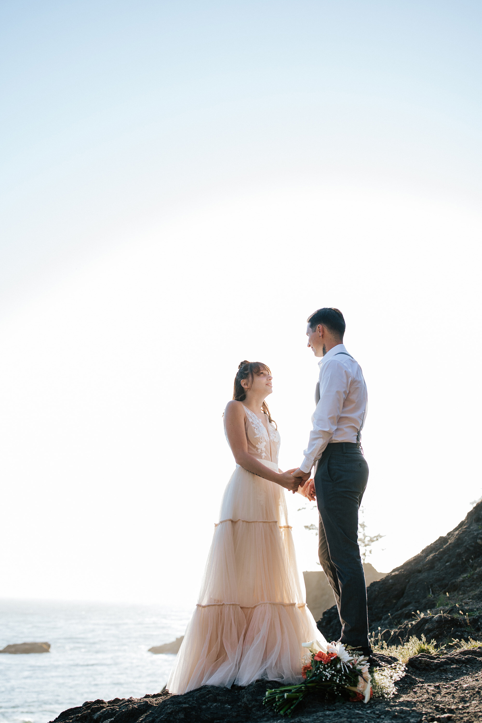  Elopement photographer Emily Jenkins Photography captures a woman in a blush pink wedding gown and a man in black pants and suspenders holding hand on the Oregon Coast during an elopement session. elopement outfit inspiration blush pink wedding dres