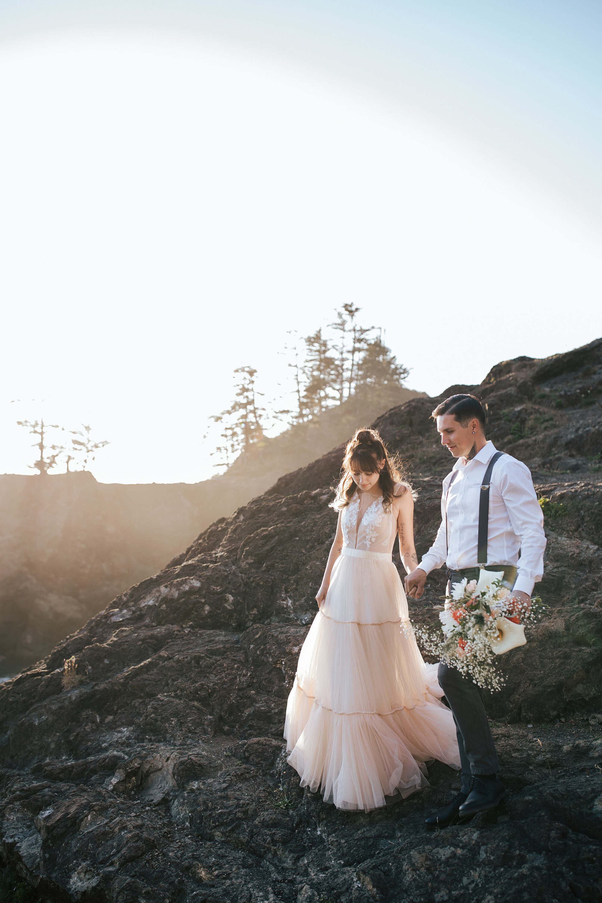  During a destination elopement a stylish couple wearing blush gown and black suspenders walk down a cliff on the coast of brookings, Oregon during sunrise by Emily Jenkins Photography. sunrise elopement shots intimate wedding elopement ideas #emilyj