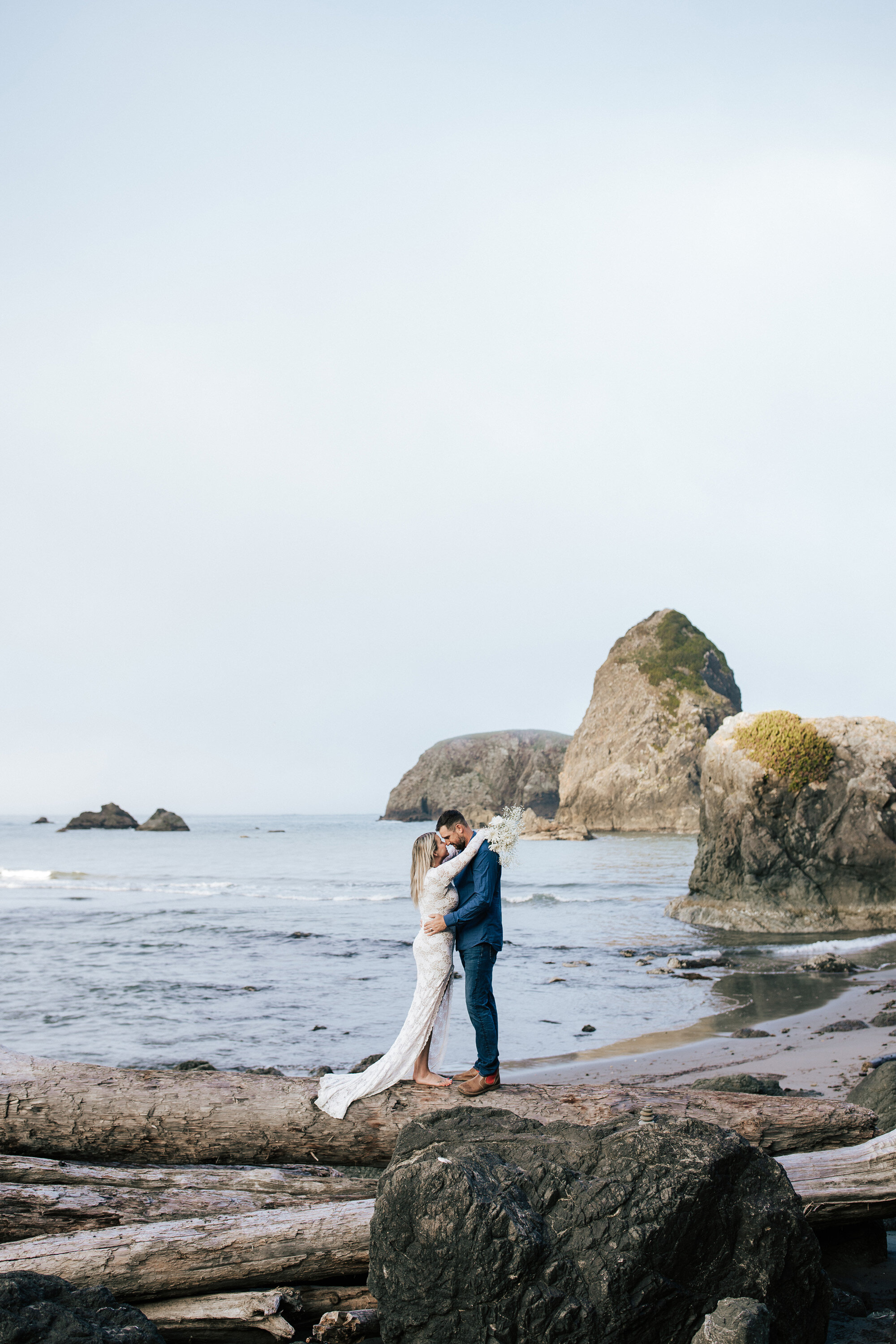  On a giant log on the Brookings, Oregon beach a woman has her arms around her husband’s neck going in for a kiss taken by elopement photographer Emily Jenkins Photography. couple poses elopement photography ideas USA west coast elopement locations #