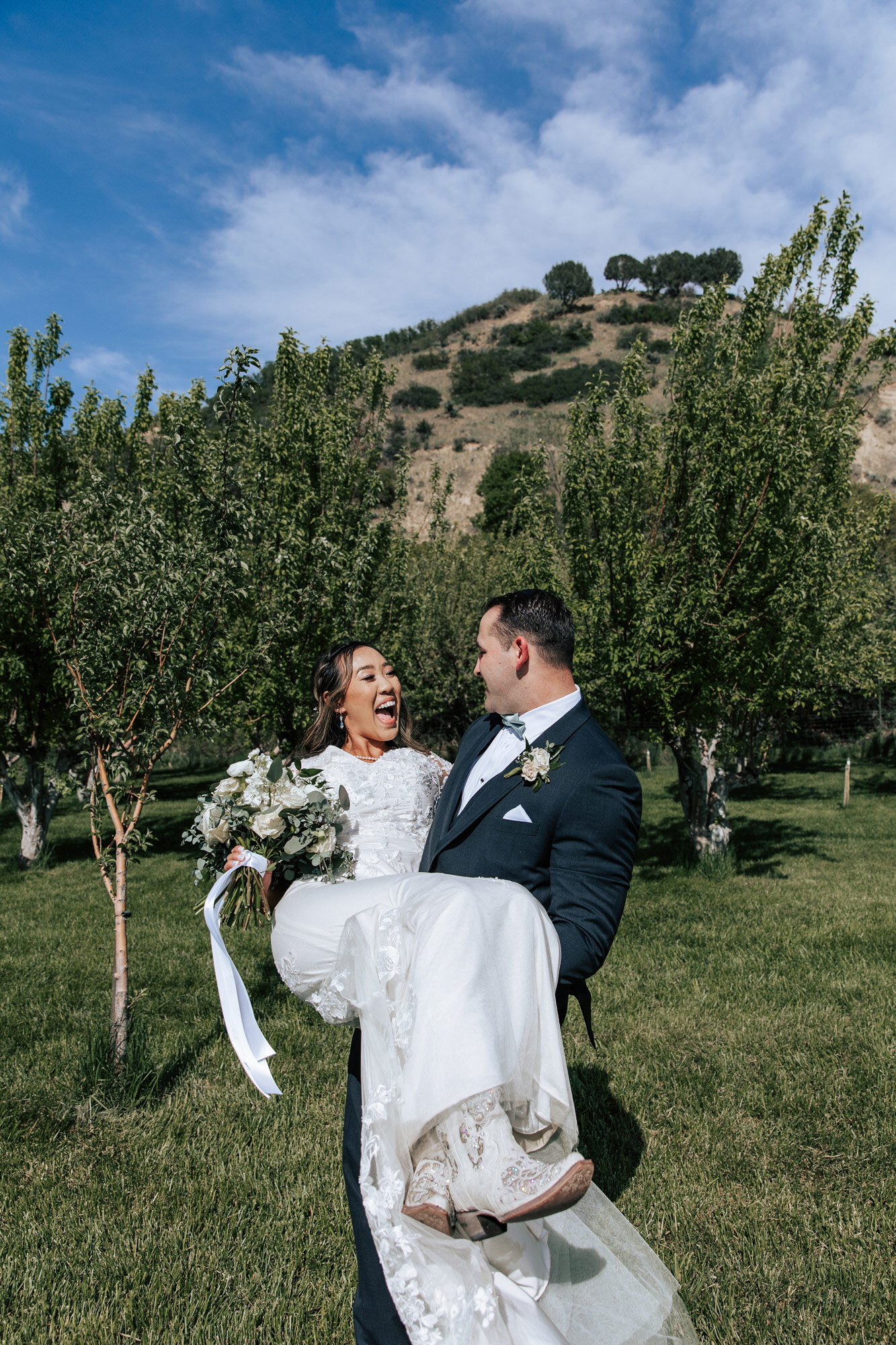  Emily Jenkins Photography captures the groom holding his bride in his arms as she laughs happily at him in front of an orchard in Mapleton, Utah. white wedding cowgirl boots boots and dress #emilyjenkinsphotography #emilyjenkinsweddings #utahwedding