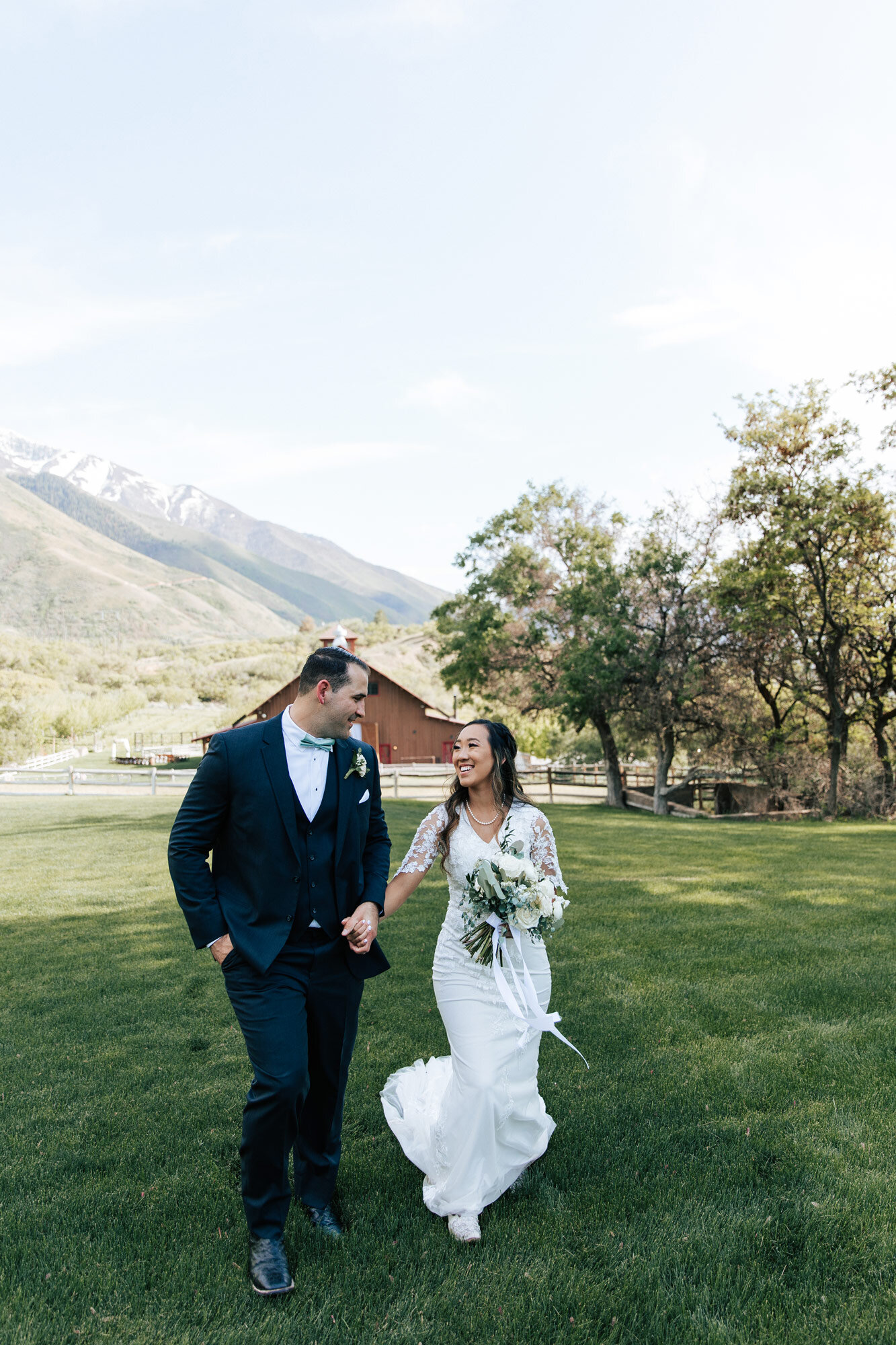  At Quiet Meadow Farms a bride with white cowgirl boots looks lovingly at the groom as they walk in a green meadow in front of a barn by Emily Jenkins Photography. barn and blue sky wedding green bow tie groom #emilyjenkinsphotography #emilyjenkinswe