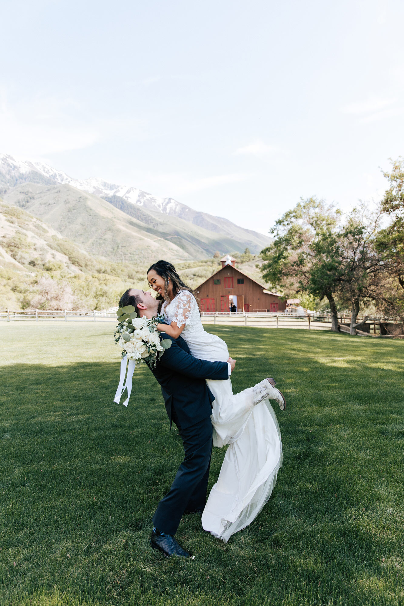  With blue sky and brown barn behind a groom holds his bride up in his arms as she goes in for a kiss in Utah County by Emily Jenkins Photography. blue sky wedding photos country summer wedding in Utah #emilyjenkinsphotography #emilyjenkinsweddings #