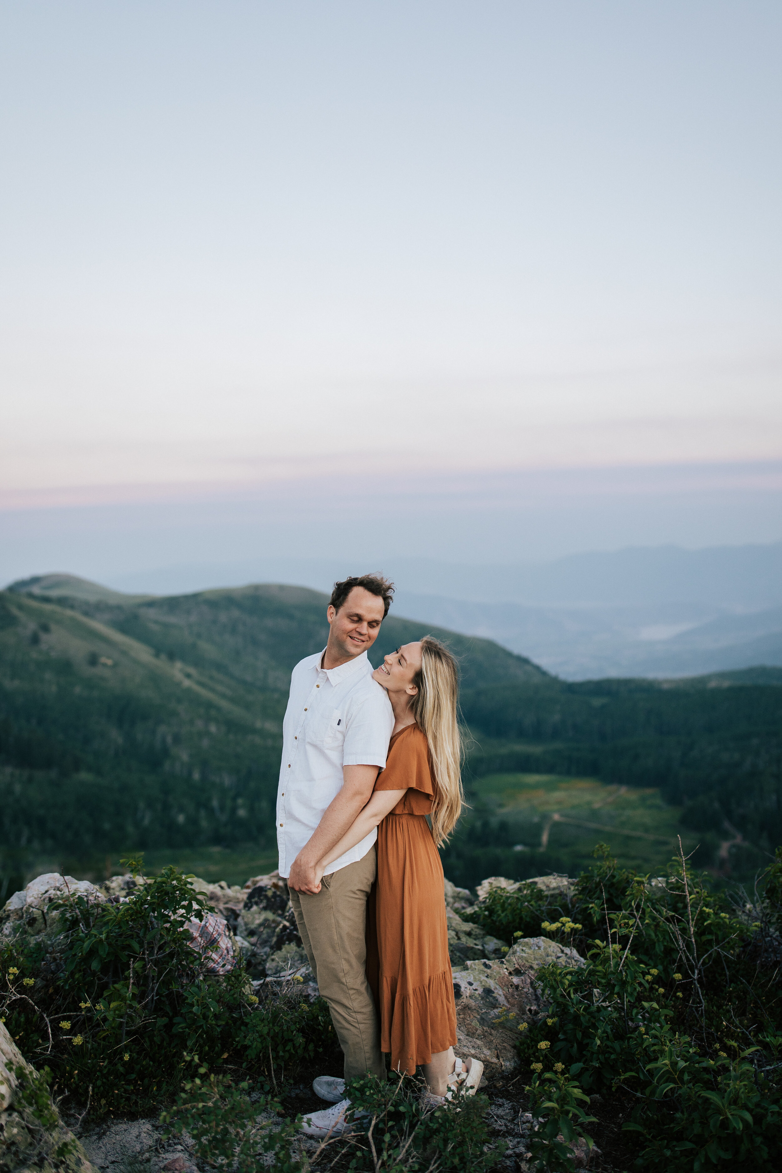  Engagement session in wildflowers in the mountains near Park City, Utah. Summertime engagement photos with mountain views. Engaged couple doing a piggyback ride in the mountains. Outfit inspo. Engagement outfits. Rocky mountain cliffs with pine tree