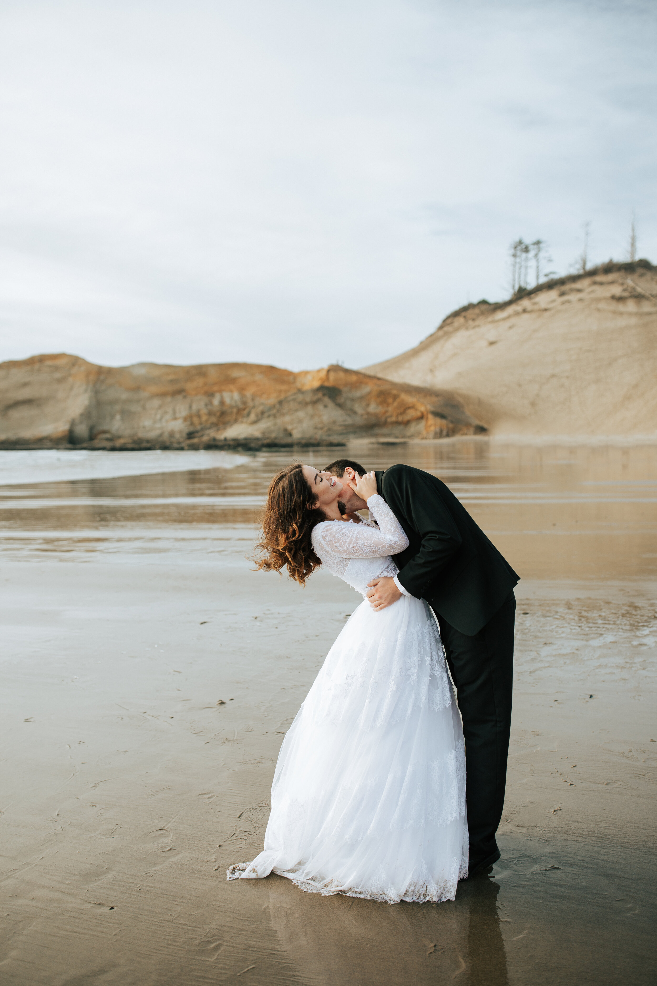  Emily Jenkins Photography captures a husband kissing a bride's neck on Cannon Beach, Oregon during an elopement. intimate wedding photography big beautiful beach for wedding photography white wedding gown and black suit #emilyjenkinsphotography #emi