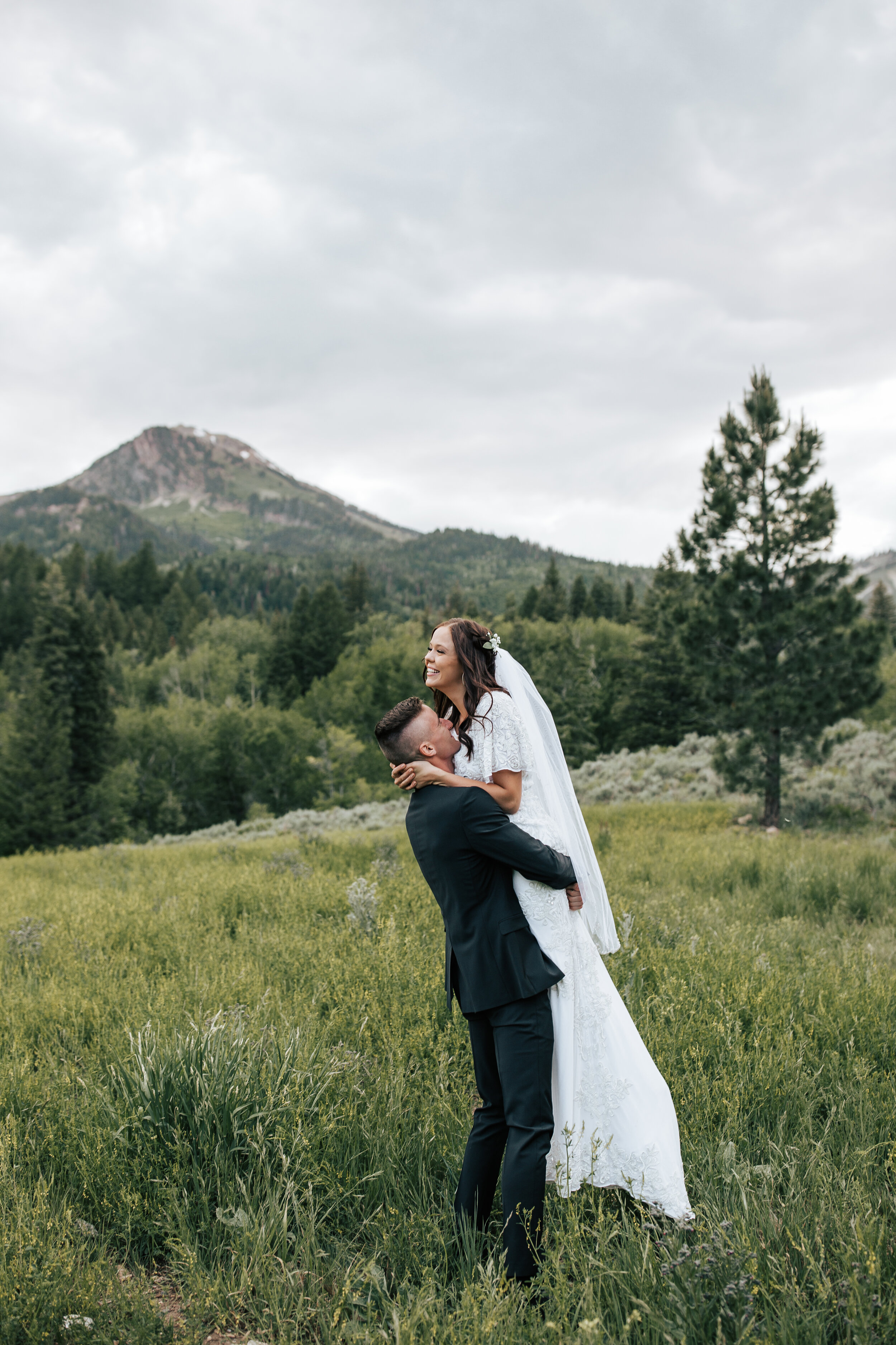  A groom holds his laughing bride up in his arms in a green grass field in the summertime in the Utah Mountains by Emily Jenkins Photography. Utah mountain elopements stunning elopement photography groom holding bride #emilyjenkinsphotography #emilyj