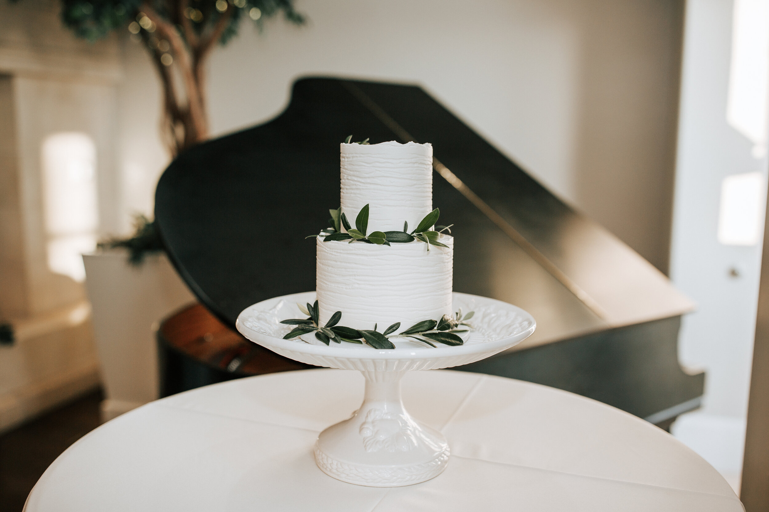  At a wedding venue in Orem, Utah Emily Jenkins Photography captures a photo of a two tier simple and class wedding cake with greenery around the sides for embellishments. simple classy wedding cake two tier wedding cake white cake #emilyjenkinsphoto