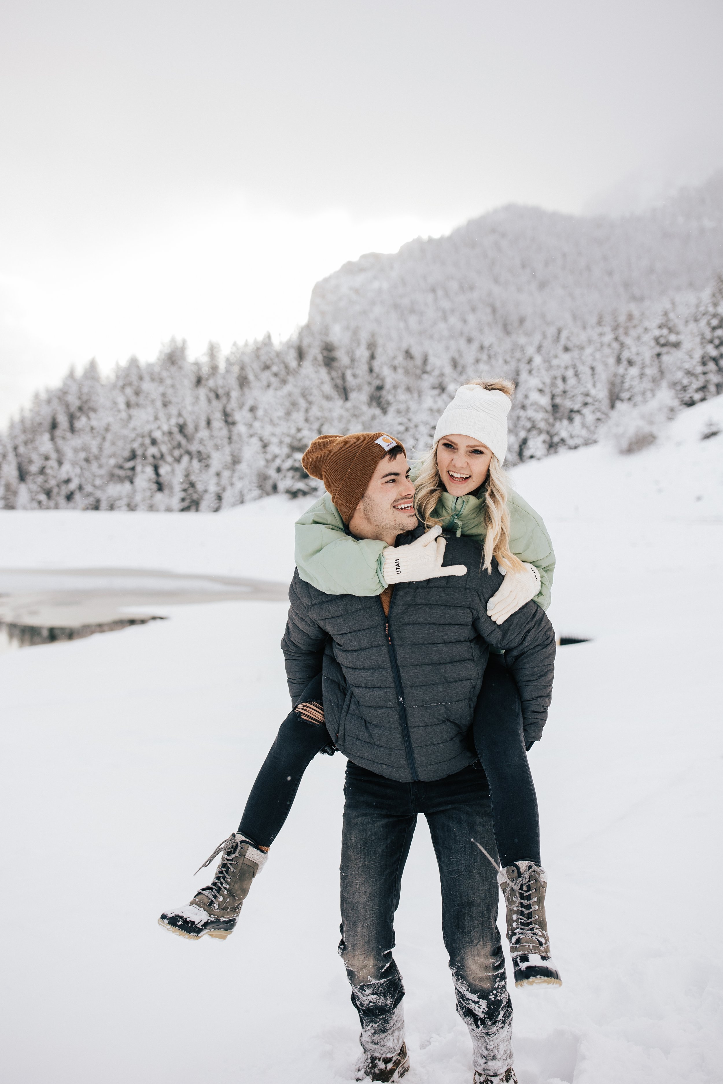 Tibble Fork winter engagement session winter outfit inspo coats and beanies man gives wife piggyback ride in the mountains