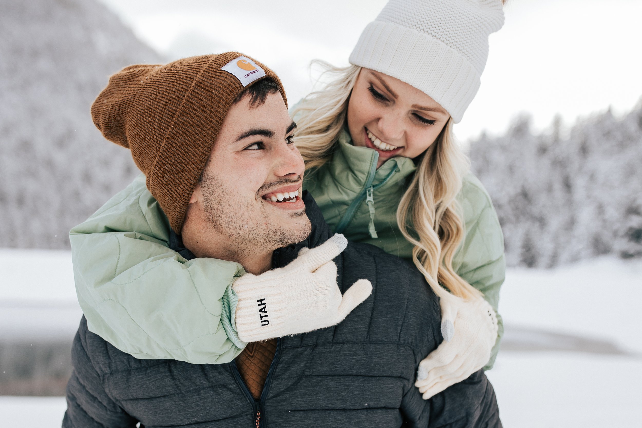 Winter engagement shoot couple wearing hats coats gloves playing in snow guy gives his fiance piggy back ride in the snow Park City Utah photographer #parkcityphotographer #utahphotographer