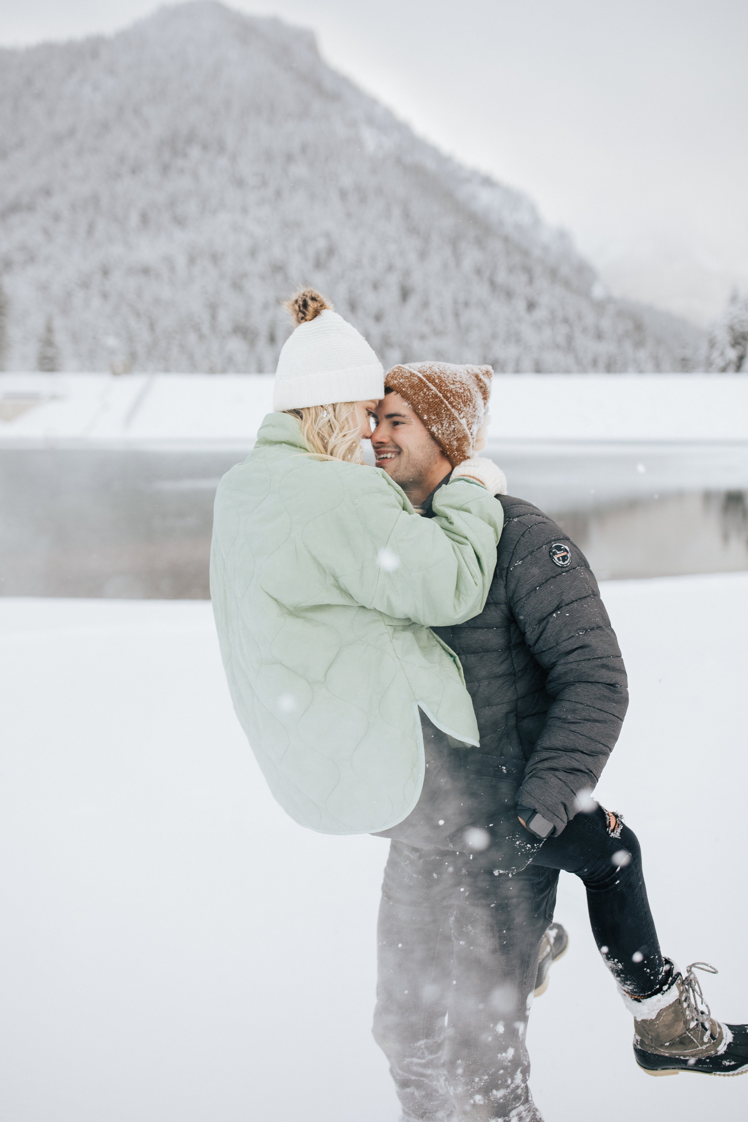  Winter photoshoot outfit inspo man holds his fiance in the mountains around pine trees and snow couples session couple playing in the snow winter engagement session couple wear coats and beanies with winter coats in Park City #winteroutfitinspo #out