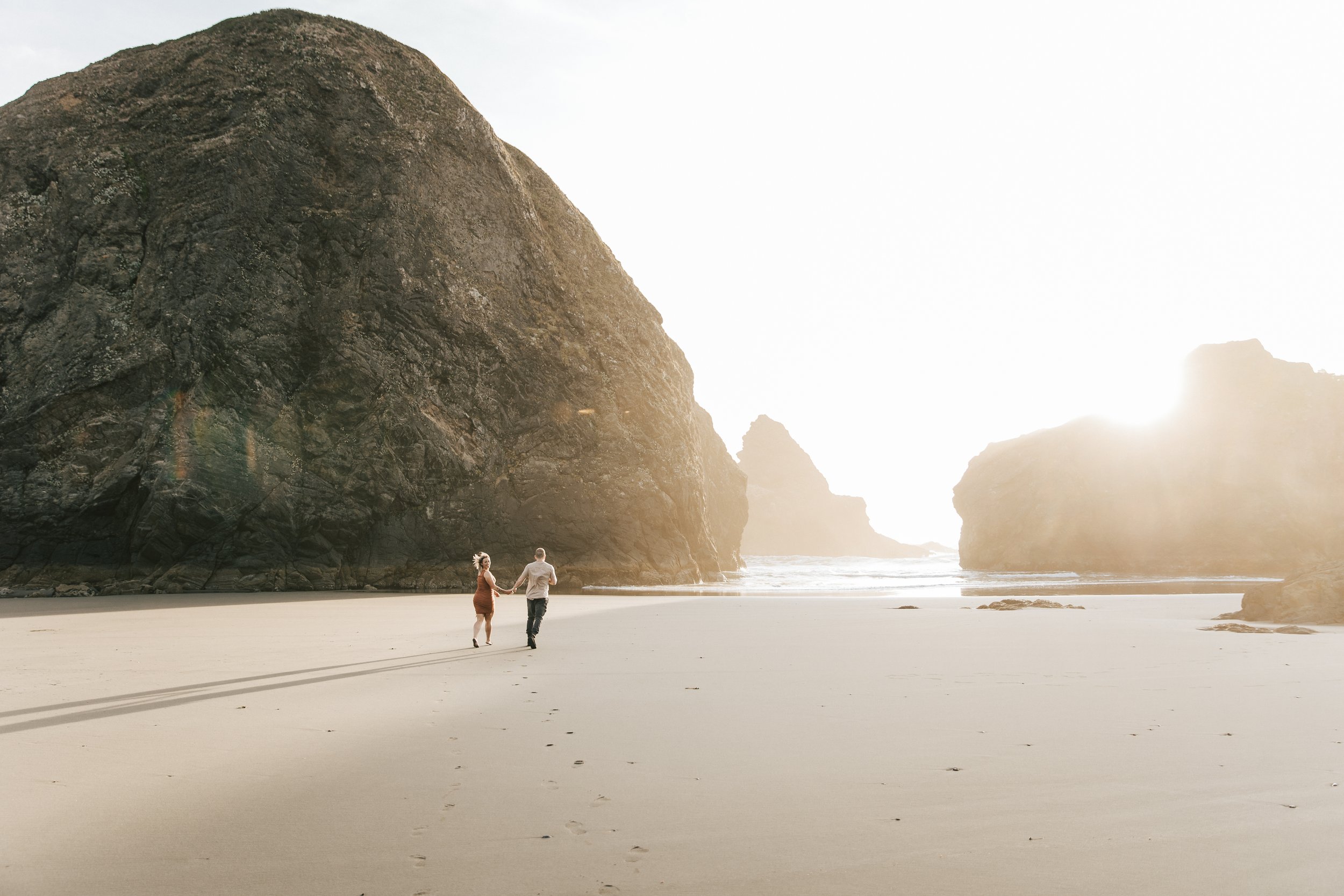  Couple running along the beach at golden hour on the southern Oregon coast with the sun setting behind them. Engagement session on the Oregon coast, with wind in their hair, running away towards the water. In love couple engagement photoshoot. Emily