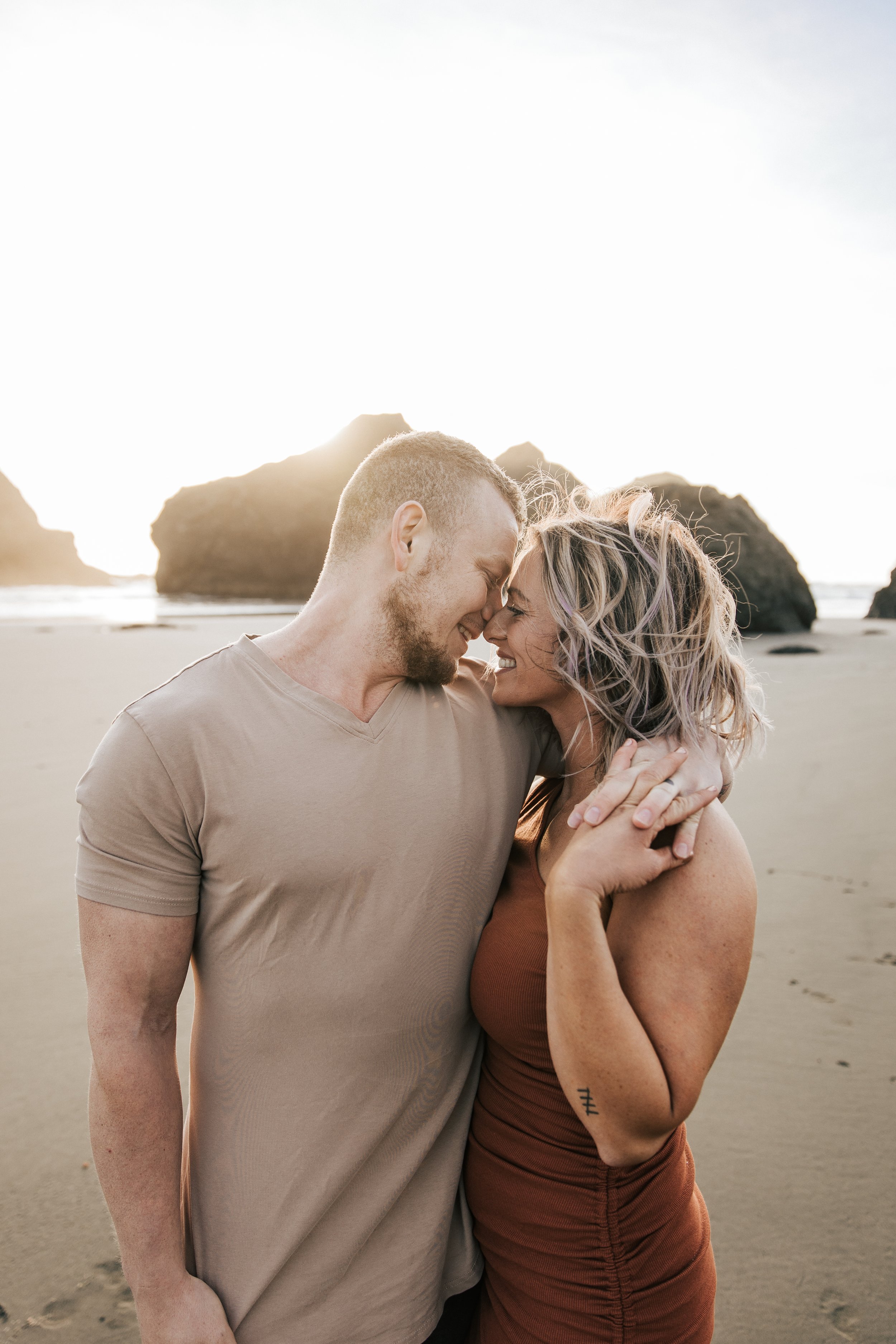  Brookings, Oregon engagement session. Couple hugs on the beach for a summer couples session on their anniversary. Husband and wife run around the beach with seastacks and rocks on the beach around them, barefoot in the sand.  