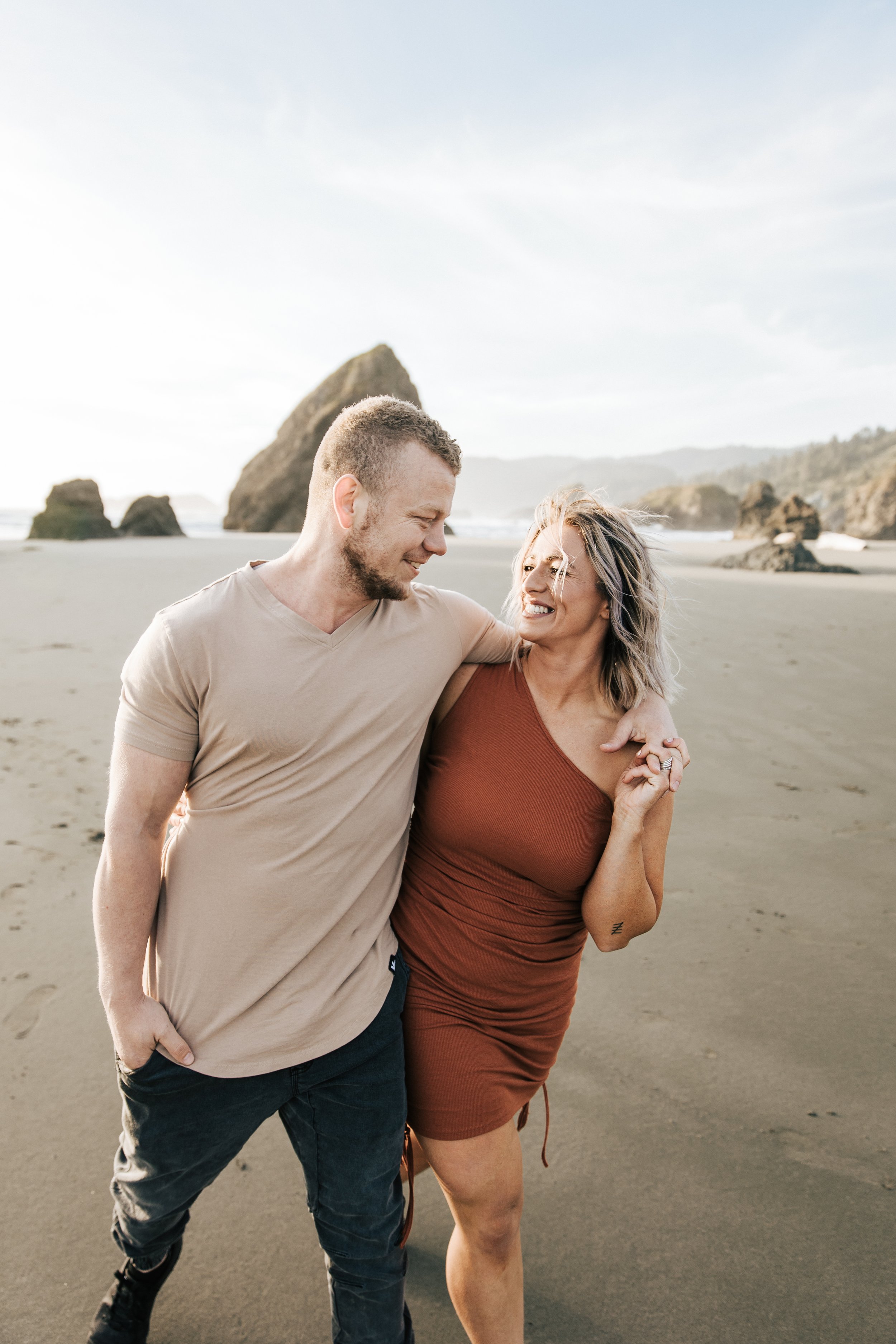  Brookings, Oregon engagement session. Couple hugs on the beach for a summer couples session on their anniversary. Husband and wife run around the beach with seastacks and rocks on the beach around them, barefoot in the sand.  