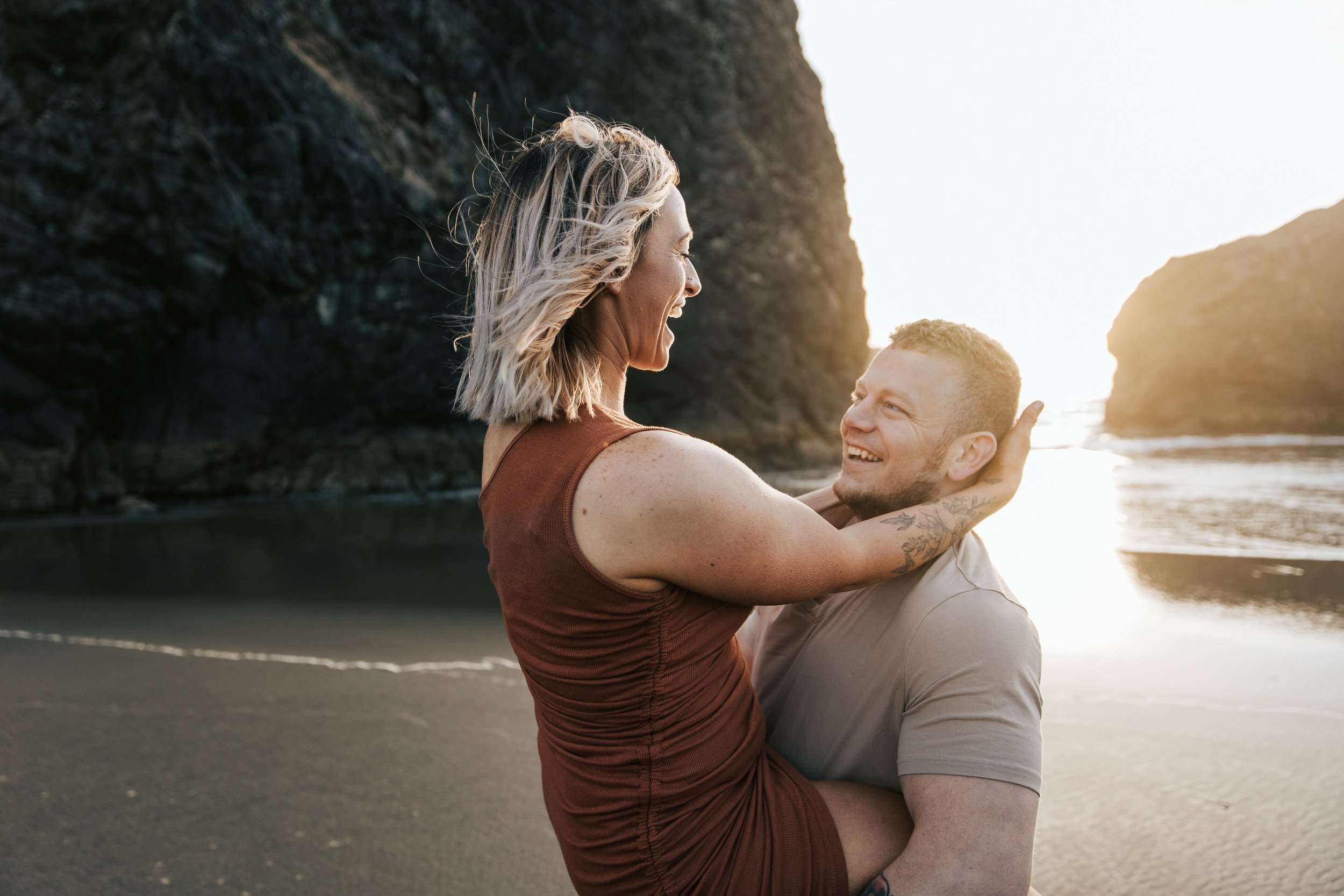  Oregon coast engagement session. Couples session in Southern Oregon. Couple playing on the beach with the sunset behind them at golden hour. Man and woman laugh together during an engagement shoot. 