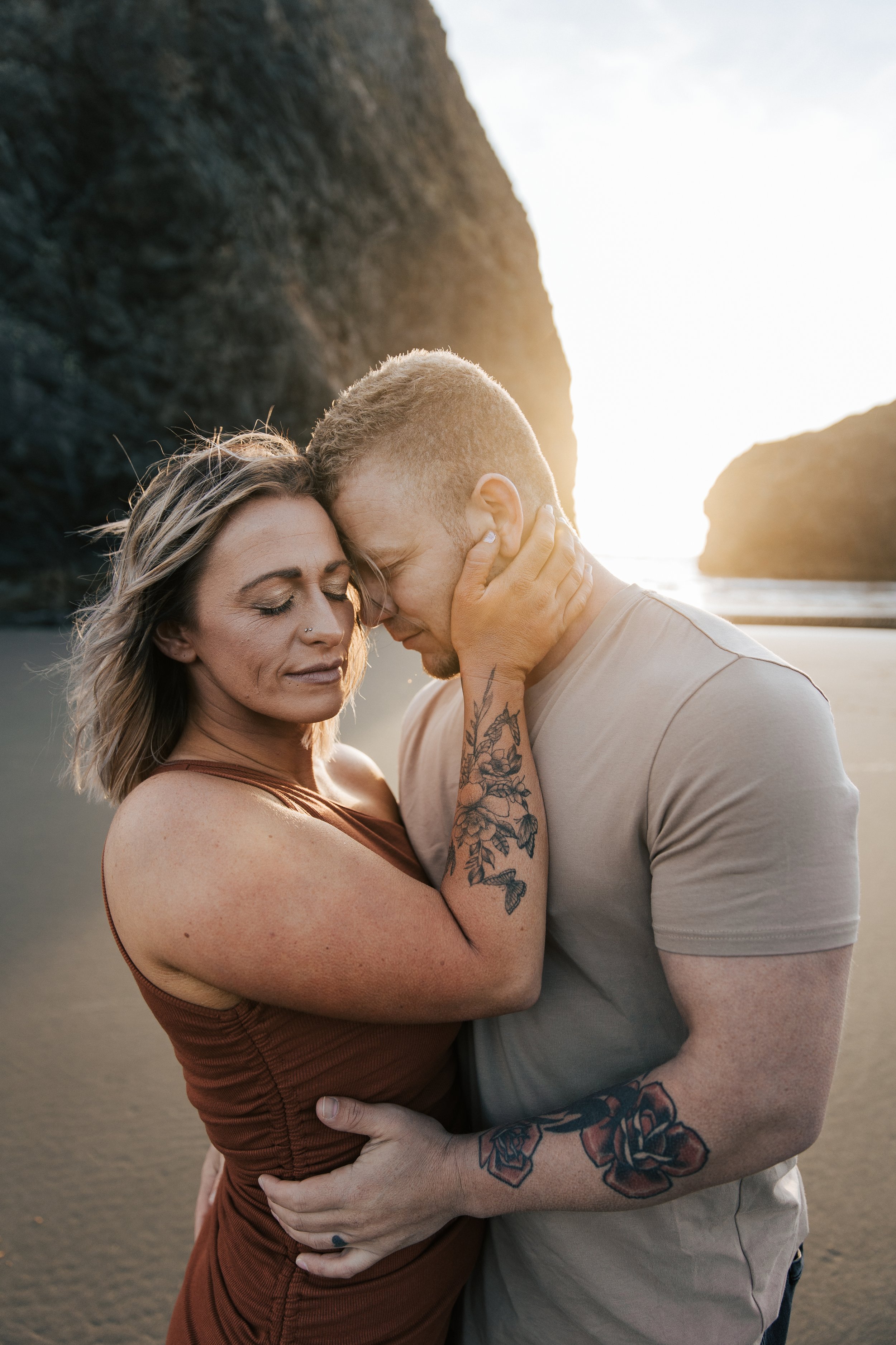  Oregon coast engagement session. Couples session in Southern Oregon. Couple playing on the beach with the sunset behind them at golden hour. Man and woman cuddle up together as the sun sets behind them between the huge rocks on the beach. 
