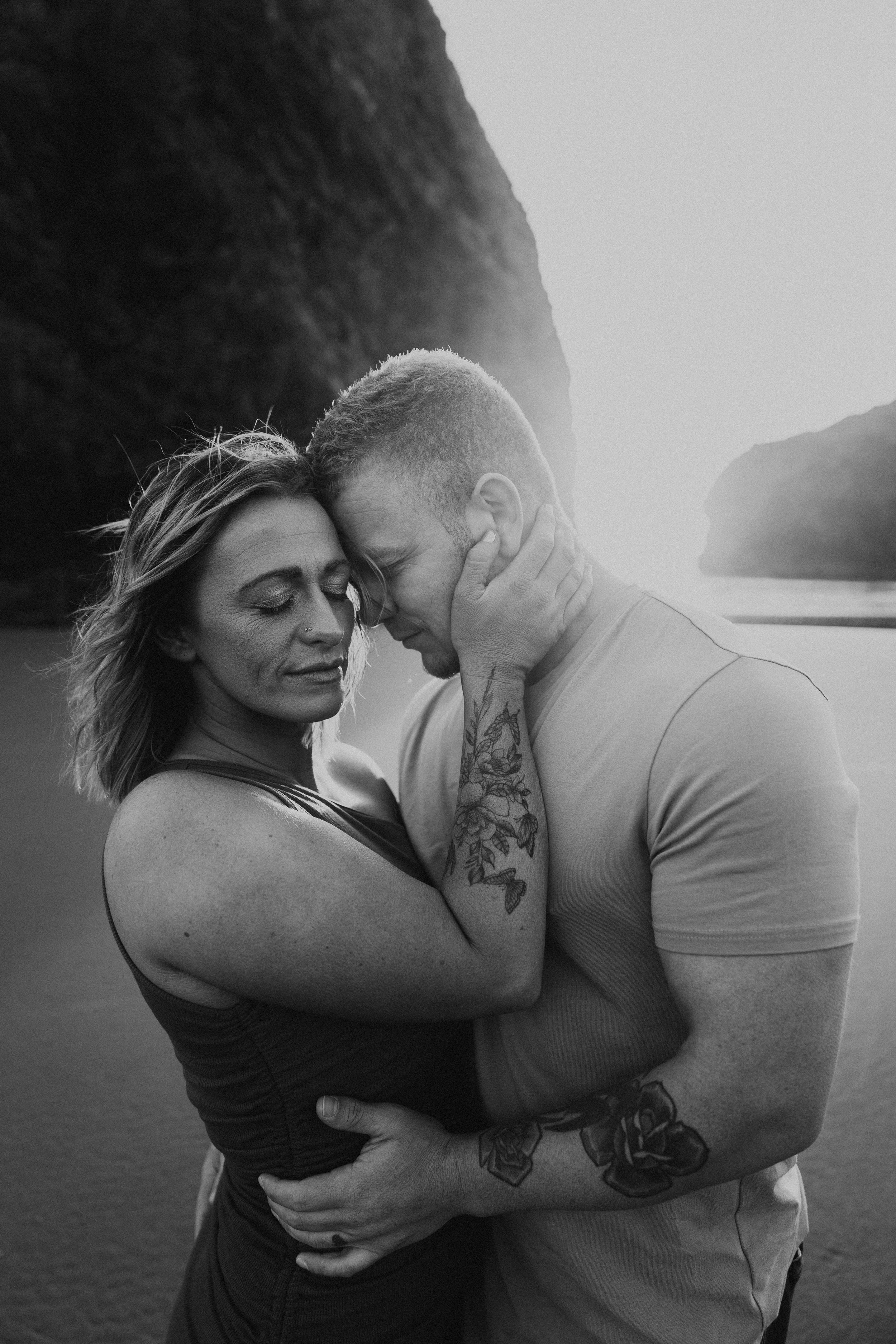  Oregon coast engagement session. Couples session in Southern Oregon. Couple playing on the beach with the sunset behind them at golden hour. Man and woman cuddle up together as the sun sets behind them between the huge rocks on the beach. Black and 