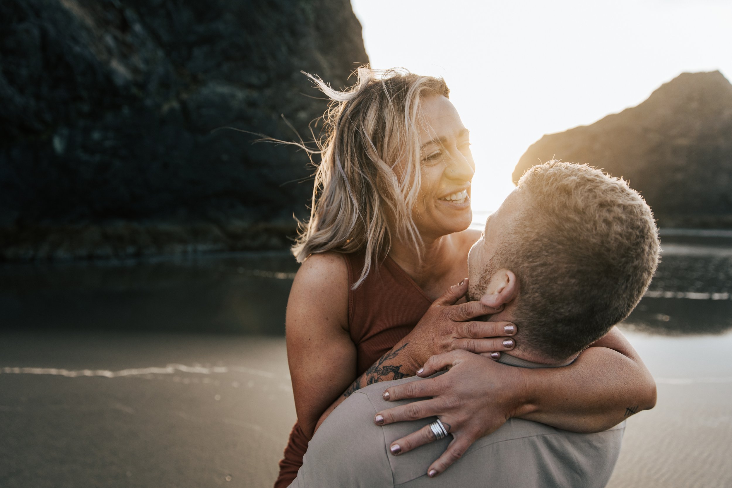  Oregon coast engagement session. Couples session in Southern Oregon. Couple playing on the beach with the sunset behind them at golden hour. Man and woman cuddle up together as the sun sets behind them between the huge rocks on the beach.   