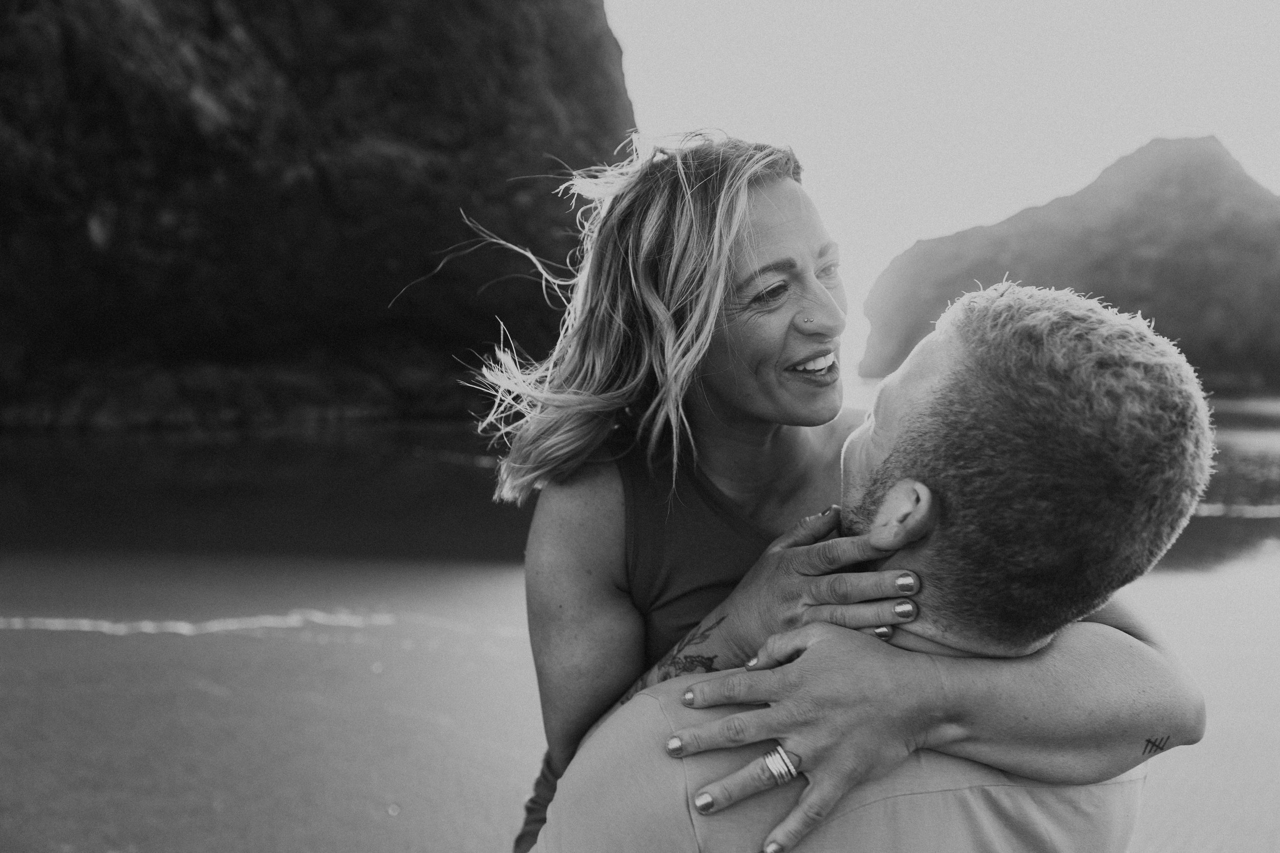  Oregon coast engagement session. Couples session in Southern Oregon. Couple playing on the beach with the sunset behind them at golden hour. Man and woman cuddle up together as the sun sets behind them between the huge rocks on the beach. Black and 
