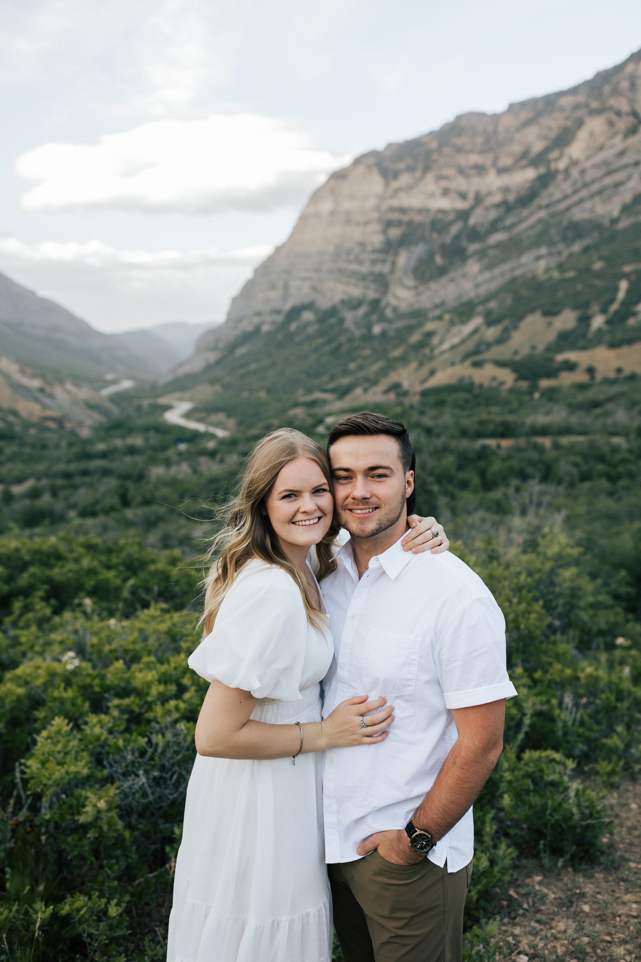  Summer anniversary couples session in the mountains. A young couple smiles at the camera in the mountains with green trees and bushes surrounding them. Utah photographer. Engagement session. #utahphotographer #utahengagements  