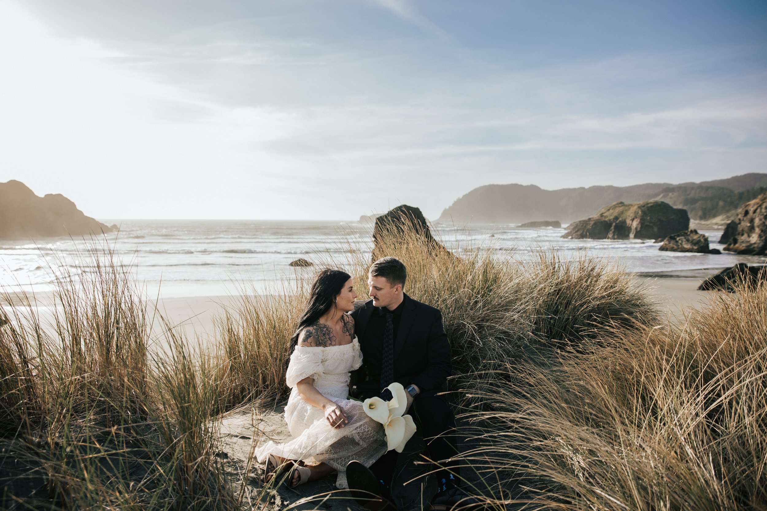  Oregon coast elopement. Couple elopes on the beach with the sun shining behind them. Sun rays behind rocks. Beach with rocks and haystacks. Southern Oregon. Beach with tall grass at golden hour. 