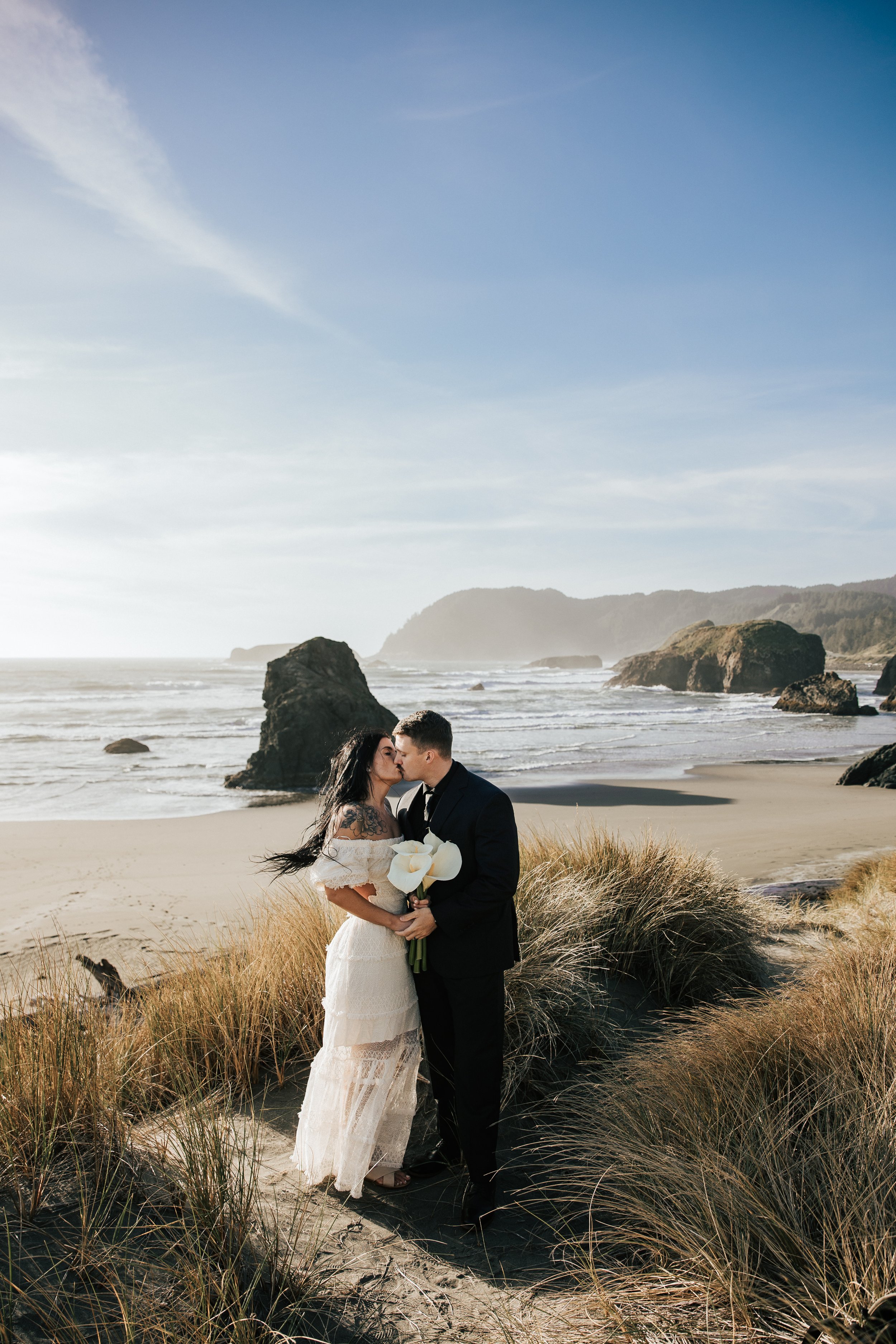  Oregon coast elopement. Couple elopes on the beach with the sun shining behind them. Sun rays behind rocks. Beach with rocks and haystacks. Southern Oregon. Beach with tall grass at golden hour. 