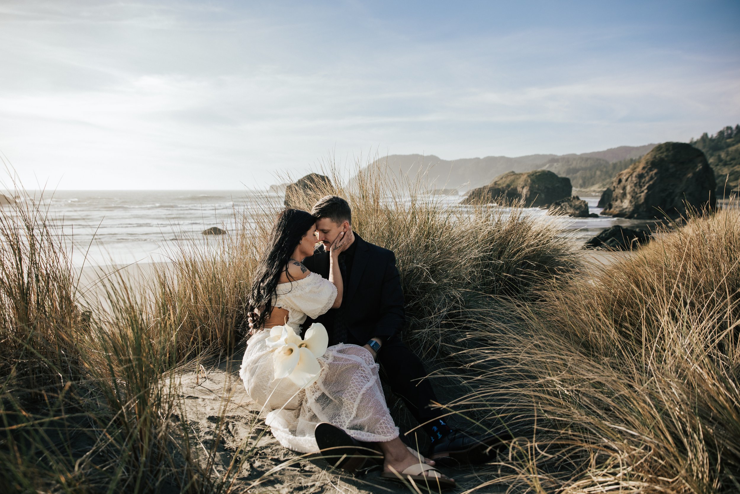  Oregon coast elopement. Couple elopes on the beach with the sun shining behind them. Sun rays behind rocks. Beach with rocks and haystacks. Southern Oregon. Beach with tall grass at golden hour. 