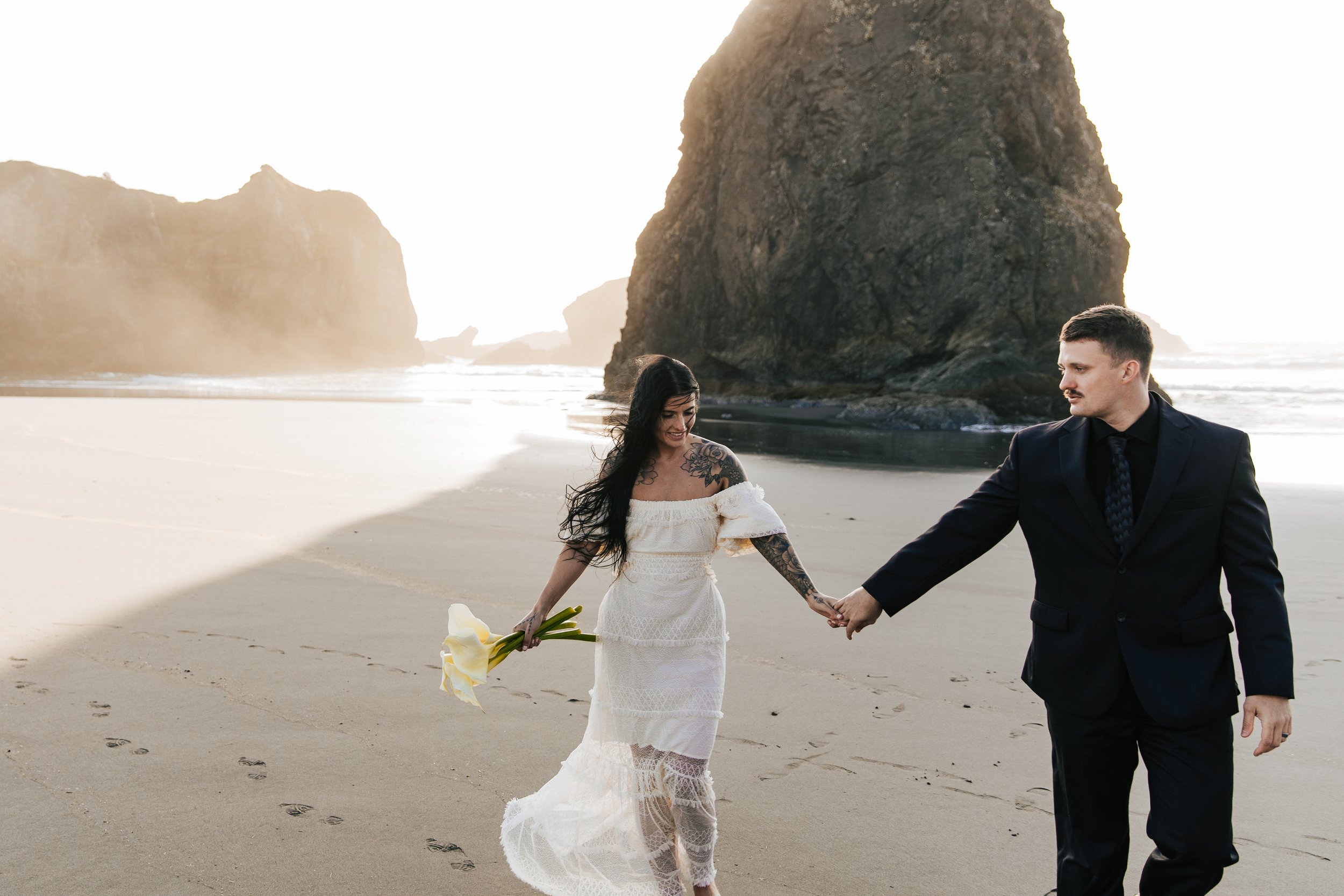  Oregon coast elopement. Couple elopes on the beach with the sun shining behind them. Sun rays behind rocks. Beach with rocks and haystacks. Southern Oregon. Couple runs together on the beach after elopement ceremony. #oregoncoast 
